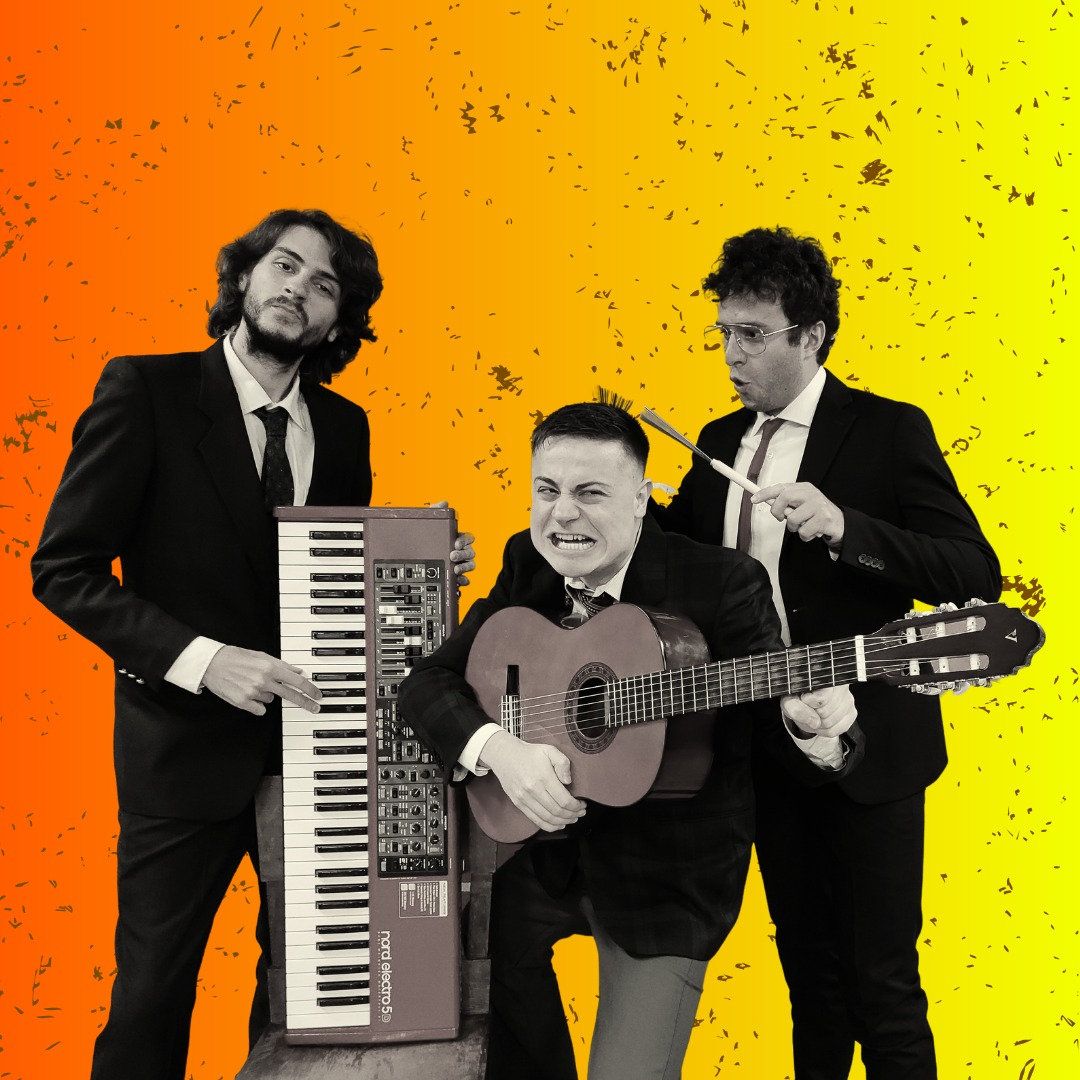 Drei Männer in Anzügen posieren vor einem leuchtend orangefarbenen und gelben Hintergrund; einer steht mit einem Keyboard, ein anderer zieht eine Grimasse, während er eine Gitarre spielt, und der dritte zeigt mit Schlagzeugstöcken auf den Gitarristen. Das Bild hat eine spielerische, energiegeladene Ausstrahlung.