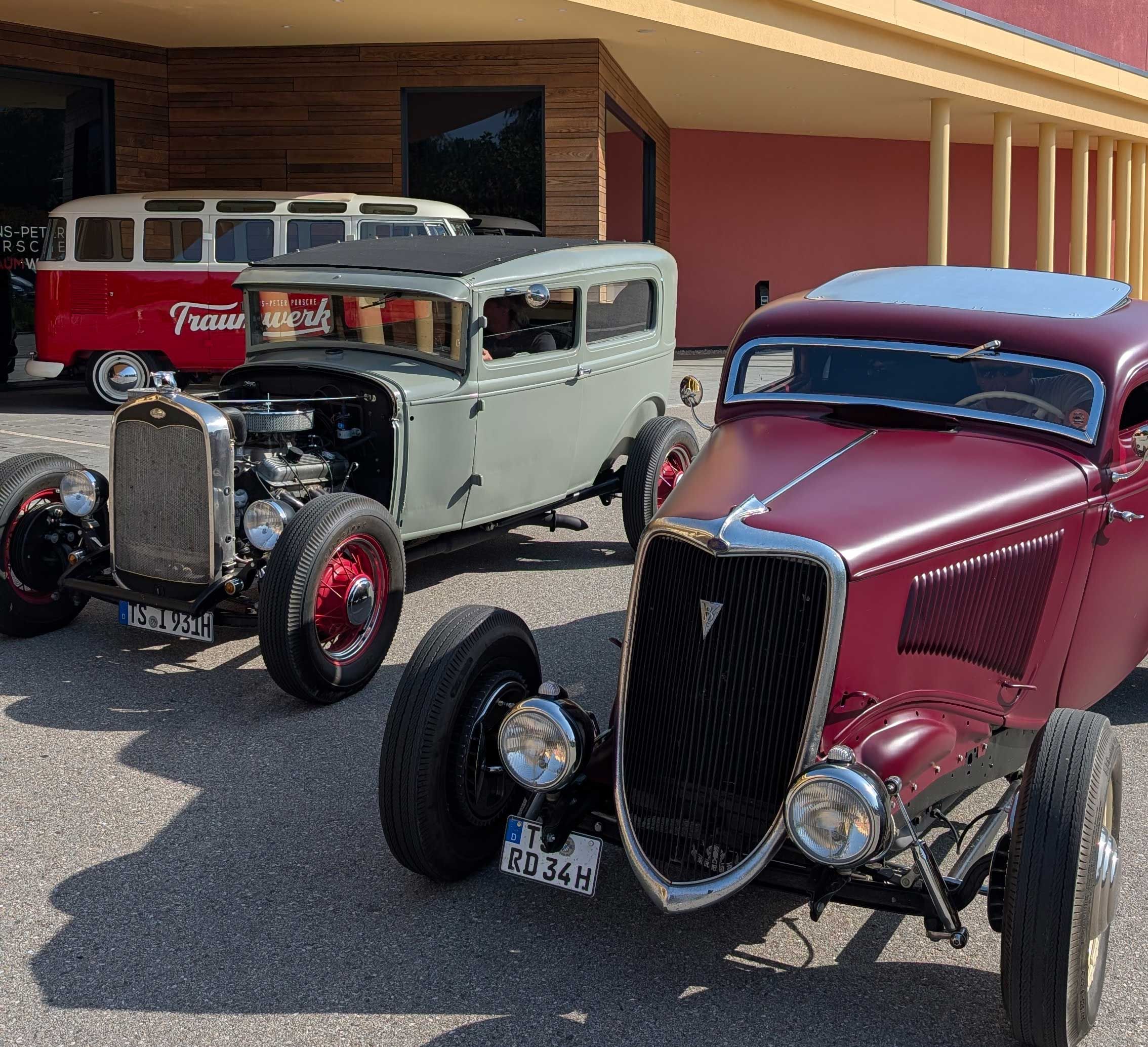 Drei Oldtimer sind vor einem Gebäude geparkt: vorne ein kastanienbrauner Hot Rod, dahinter ein grüner Oldtimer mit roten Rädern, und im Hintergrund ein weiß-roter Retro-Van.
