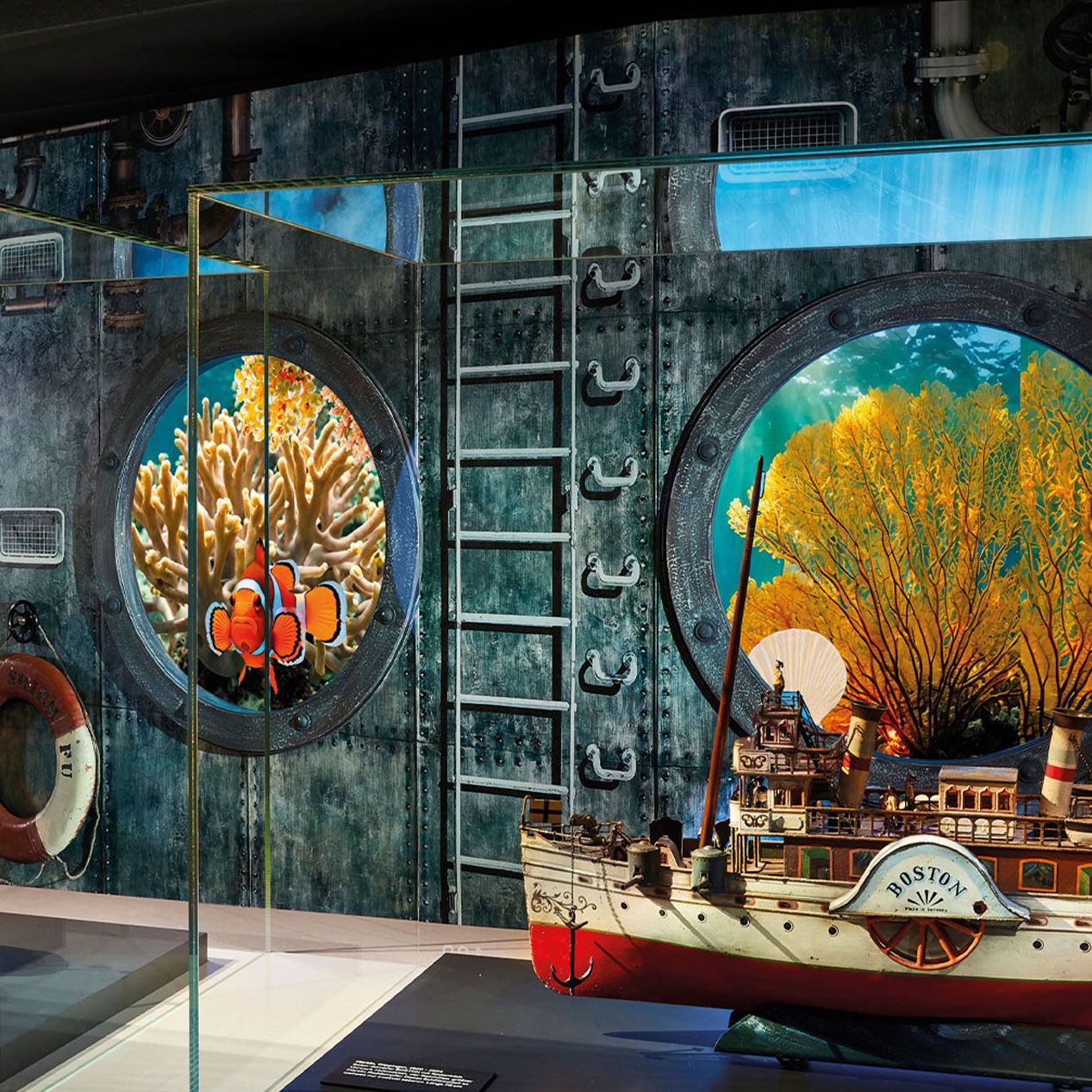 In einem Museum ist ein Schiffsmodell mit dem Namen Boston vor einer Glasscheibe ausgestellt, die Unterwasserszenen mit Korallen, einem Clownfisch und einem gelben Seefächer zeigt, vor einem Hintergrund, der an das Innere eines U-Boots mit Bullaugen und einer Leiter erinnert.
