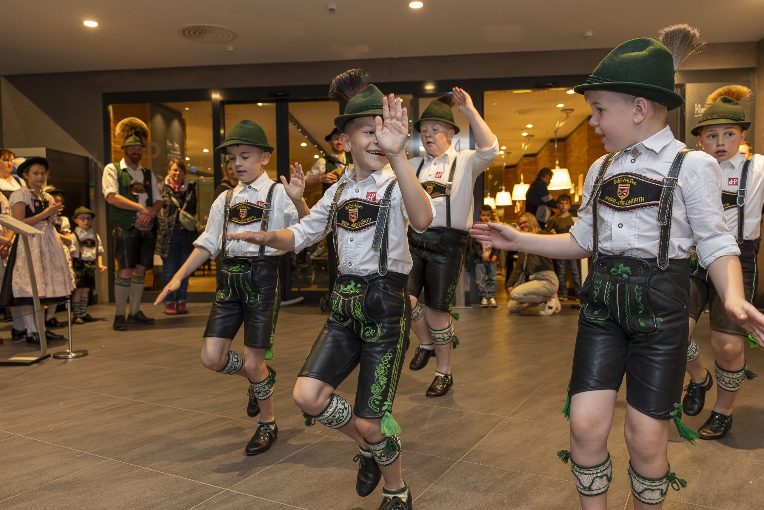 Junge Burschen in traditioneller bayerischer Tracht führen im Haus einen Volkstanz auf, heben die Hände und lächeln, während Menschen in ähnlicher Tracht im Hintergrund zusehen.