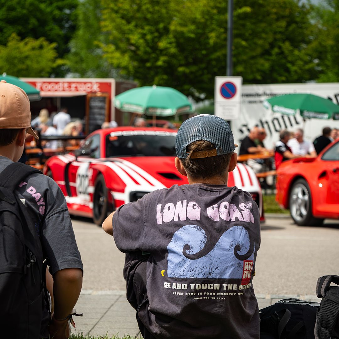 Zwei Personen mit Rucksäcken beobachten bei einer Veranstaltung im Freien ein rot-weißes Rennauto, das vorbeifährt. Im Hintergrund sind Essensstände, grüne Regenschirme und andere Zuschauer zu sehen.