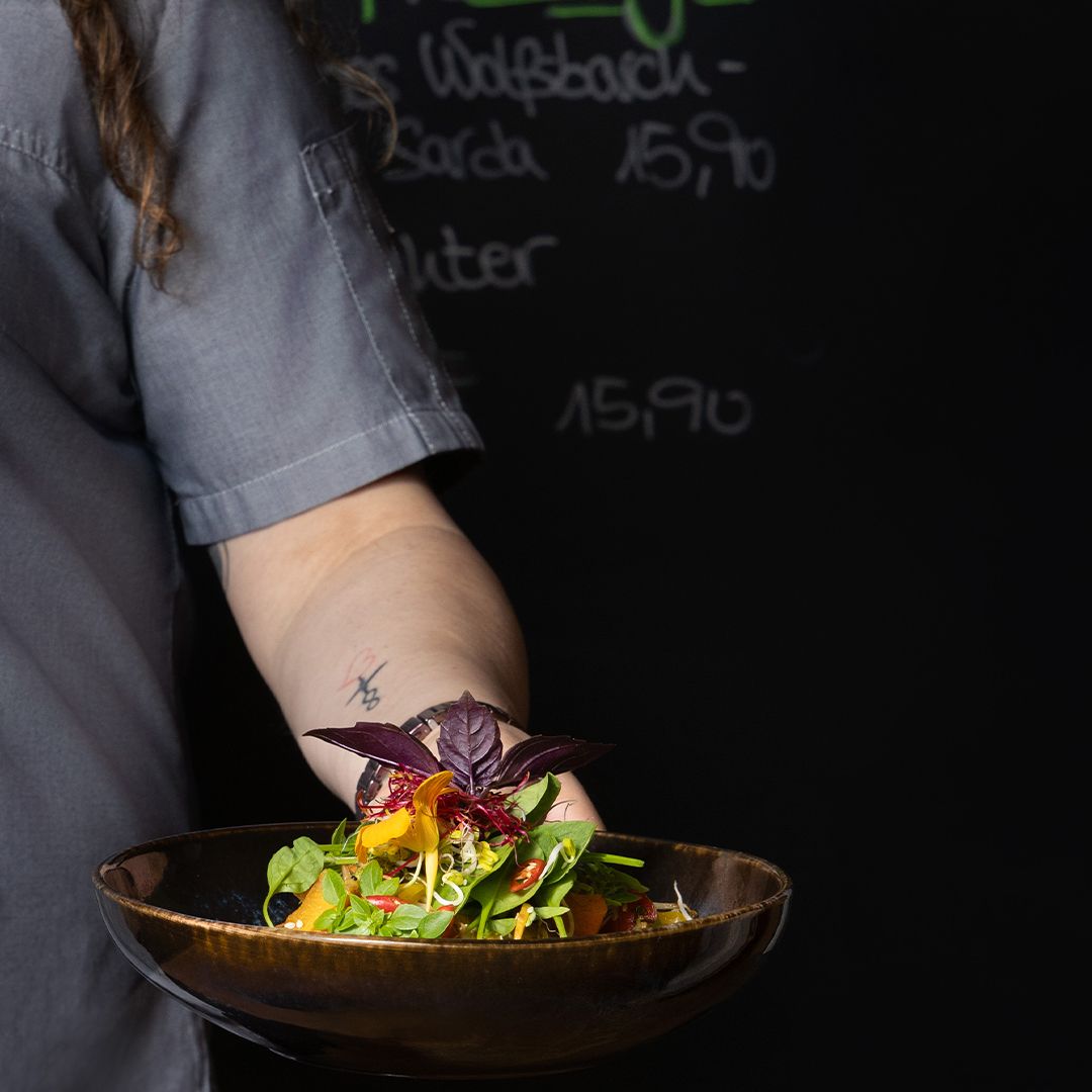 Eine Person mit langen Haaren und grauem Hemd hält eine braune Schüssel mit buntem Salat und violetten Basilikumblättern. Der Hintergrund ist schwarz, mit verschwommener Kreideschrift und Preisen.