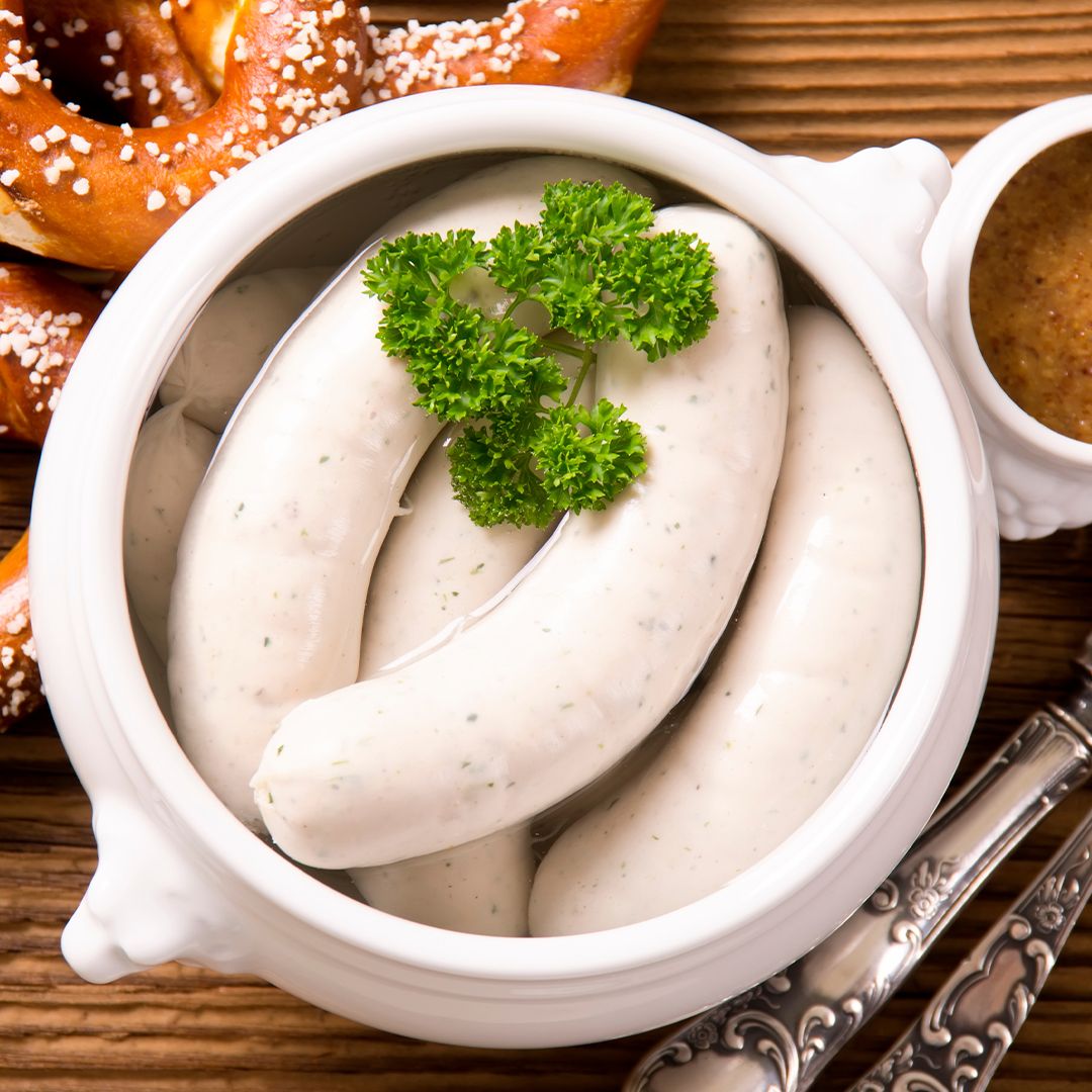 Eine weiße Schüssel gefüllt mit traditionellen Weißwürsten, garniert mit Petersilie, steht neben einer weichen Brezel und einem kleinen Schälchen Senf auf einem Holztisch.