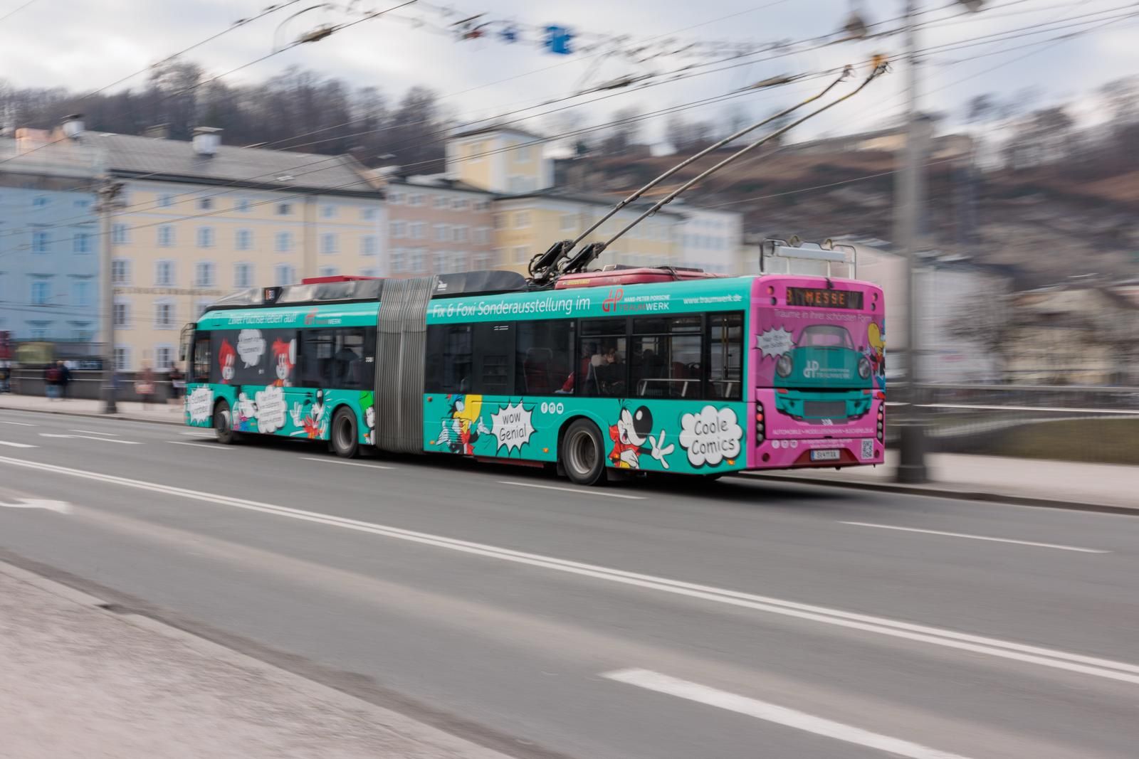 Ein bunter Gelenk-Trolleybus mit Cartoon-Grafiken fährt auf einer Stadtstraße, angeschlossen an Oberleitungen. Im Hintergrund sind unscharfe Gebäude und Bäume zu sehen, die Bewegung suggerieren.