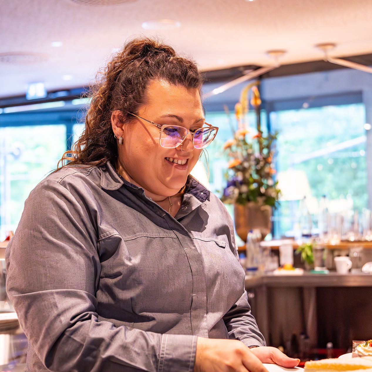 Eine Frau mit lockigem Haar und Brille, die ein graues Button-up-Hemd trägt, lächelt, während sie in einem hellen, modernen Café oder Restaurant mit Blumen und Tassen im Hintergrund arbeitet.