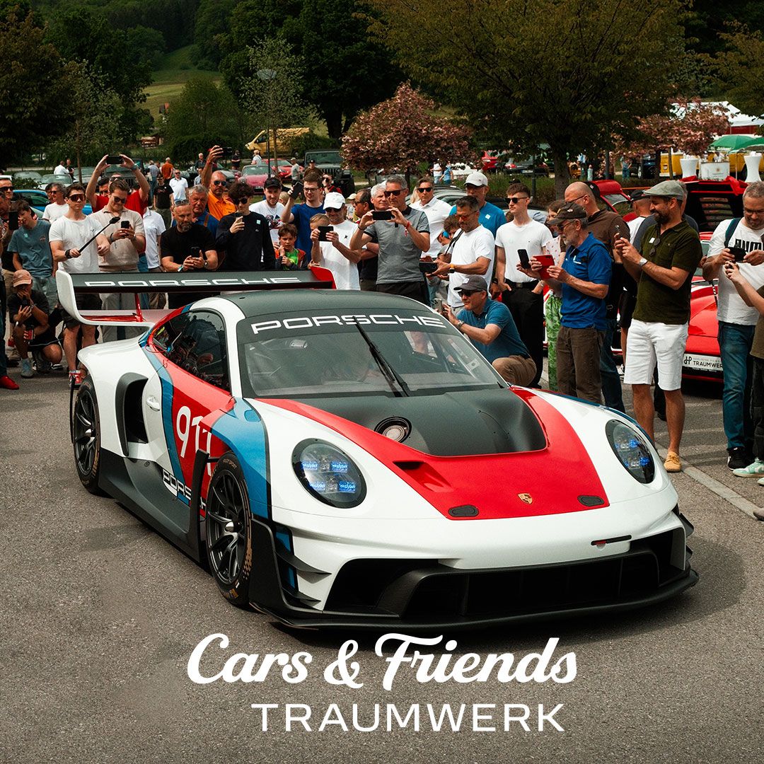 Ein Porsche-Rennwagen in rot-weiß-blauer Lackierung ist bei einem Auto-Event im Freien von einer Menge umringt, die Fotos macht. Am unteren Bildrand steht der Text „Cars & Friends TRAUMWERK“.