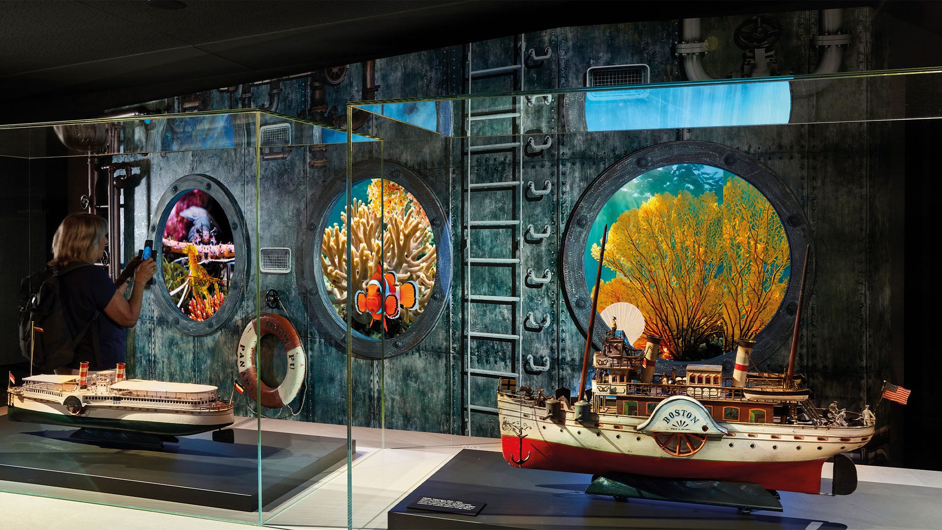Eine Person fotografiert ein Aquarium mit Bullaugen, in denen Korallen und Clownfische zu sehen sind, während im Vordergrund Modellboote in Vitrinen ausgestellt sind.