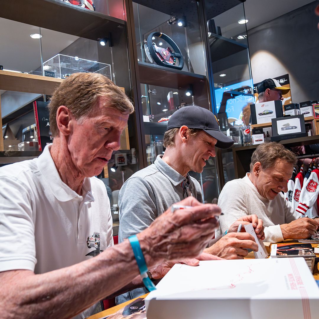 Walter Röhrl, Jörg Bergmeister und Richy Müller sitzen nebeneinander an einem Tisch und signieren Artikel in einem Laden voller Sport-Erinnerungsstücke. Sie konzentrieren sich auf Autogramme, im Hintergrund stehen Regale voller Fanartikel und Erinnerungsstücke.
