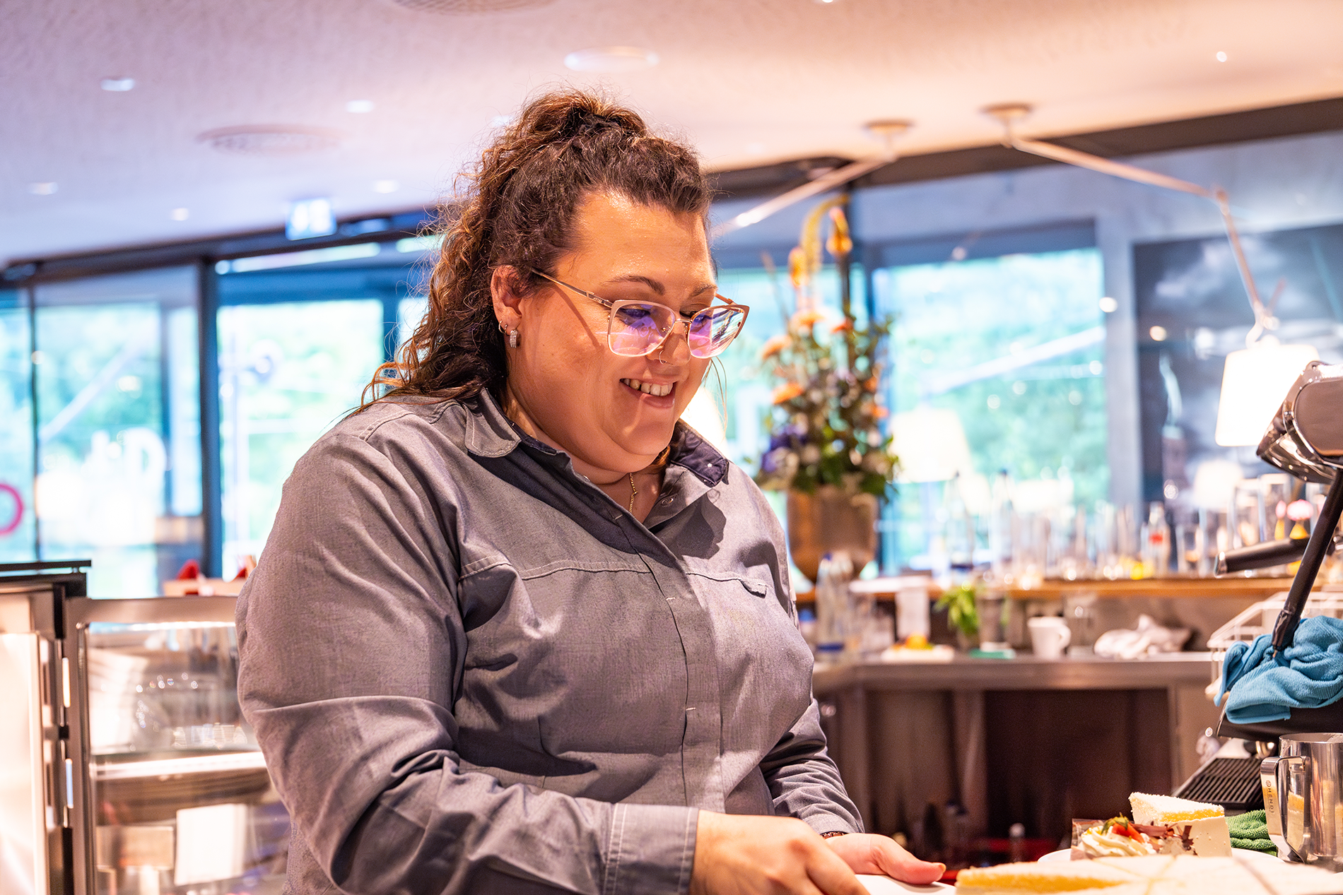 Eine lächelnde Frau mit Brille und grauem Hemd schneidet ein Stück Kuchen hinter einem Tresen in einem hellen, modernen Café mit großen Fenstern und Blumen im Hintergrund an.
