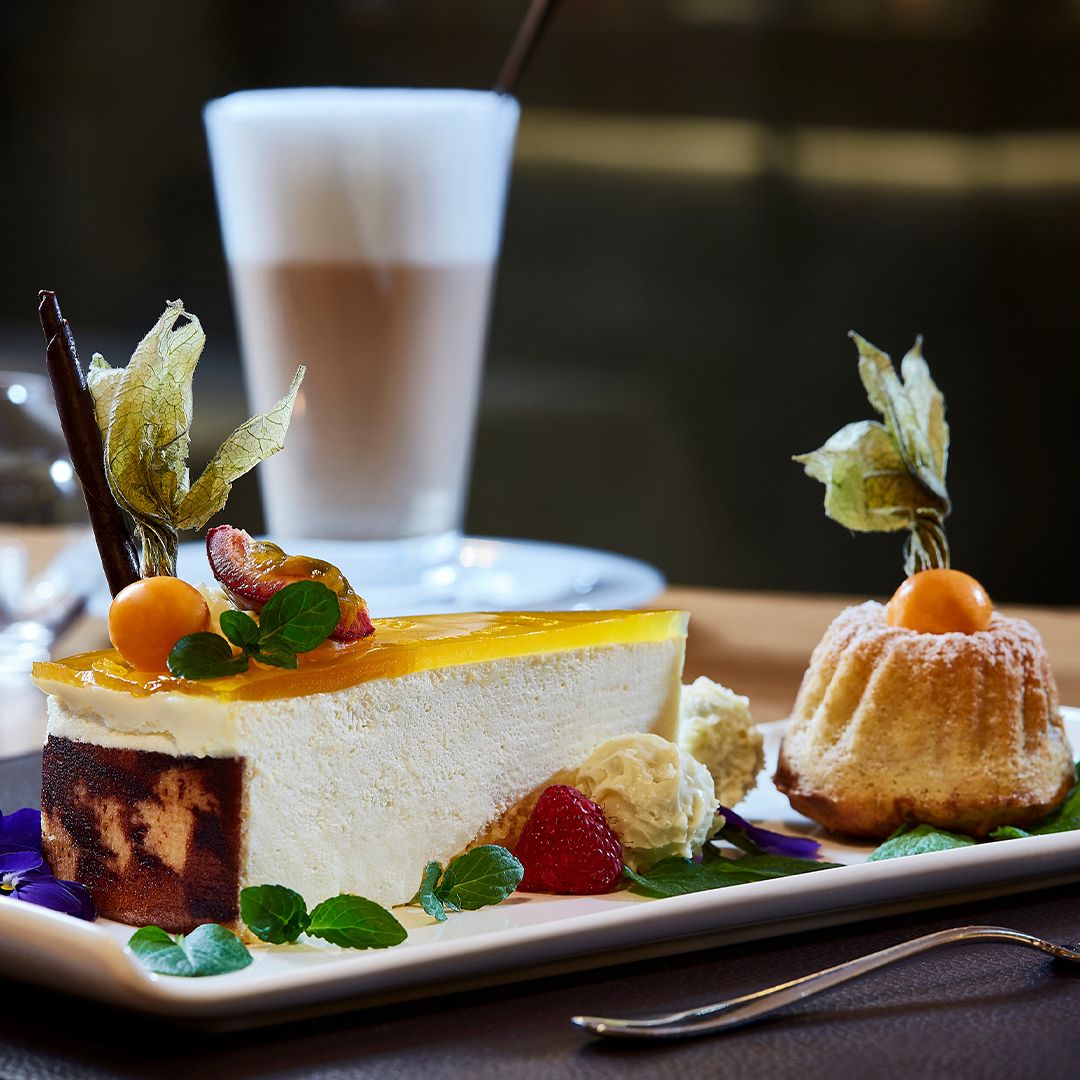 Ein Stück Käsekuchen mit Früchten, Schlagsahne und einem Schokoladenstück liegt auf einem Teller neben einem kleinen dekorierten Kuchen. Im Hintergrund ist ein Glas Latte Macchiato verschwommen. Eine Gabel liegt daneben auf dem Tisch.