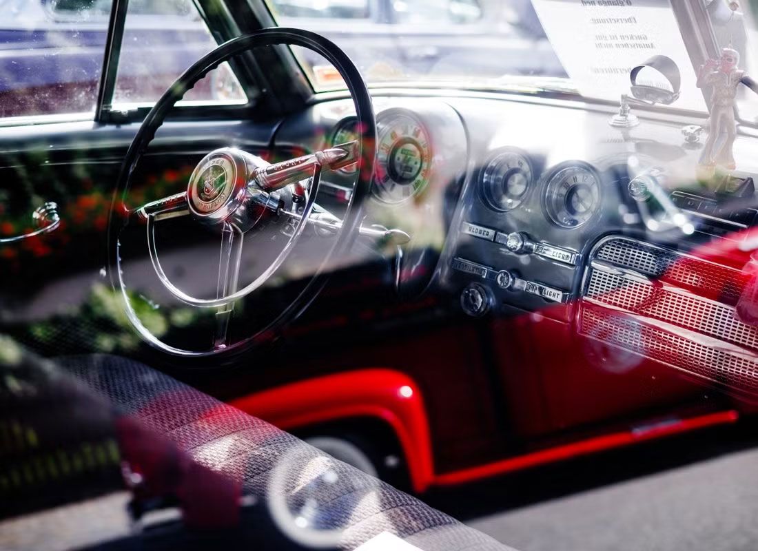 Eine Nahaufnahme des Innenraums eines Oldtimers durch das Fenster, die das Lenkrad, die klassischen Ziffernblätter des Armaturenbretts und verchromte Details zeigt, wobei die Reflexionen der Umgebung auf dem Glas sichtbar sind.