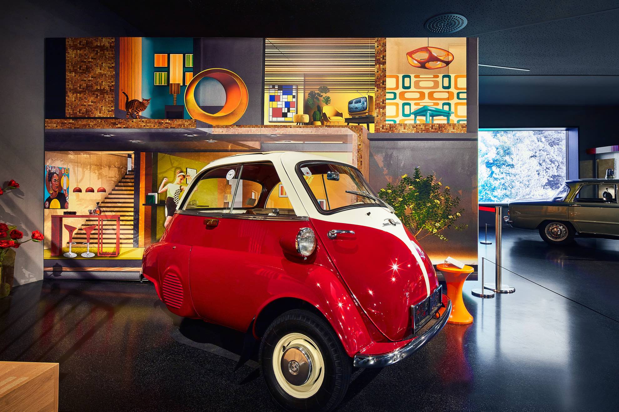Ein rot-weißer Oldtimer-Kleinwagen steht in einem modernen Museum mit lebendigem Retro-Dekor und farbenfrohen Kunstwerken im Hintergrund. Ein weiterer Oldtimer ist rechts teilweise sichtbar.