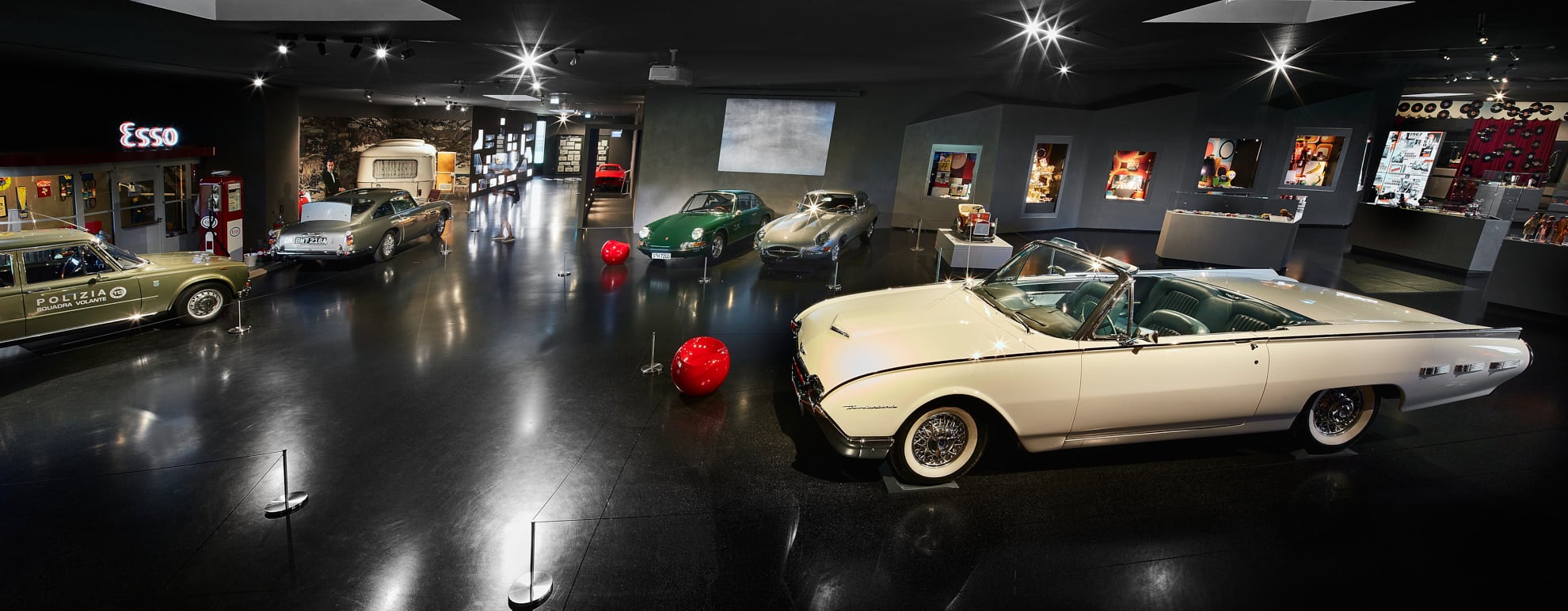 Eine geräumige, moderne Automuseumsausstellung mit Oldtimern, darunter ein weißes Cabrio im Vordergrund, ein grüner Sportwagen, ein klassisches Polizeiauto und Tankstellen-Erinnerungsstücke, alles unter sanfter Beleuchtung.