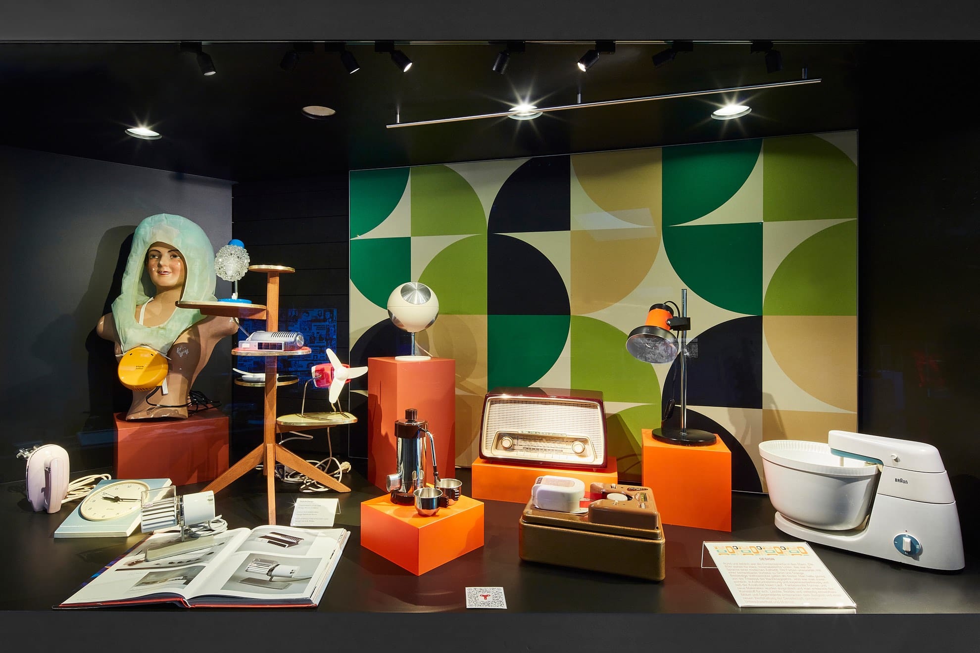 Eine Museumsausstellung mit modernen Objekten aus der Mitte des Jahrhunderts, darunter eine Schaufensterpuppenbüste, Vintage-Lampen, ein Radio, eine Toilette, Haushaltsgegenstände auf orangefarbenen Podesten und eine geometrische grün-cremefarbene Wandkulisse. Im Vordergrund steht ein offenes Buch.