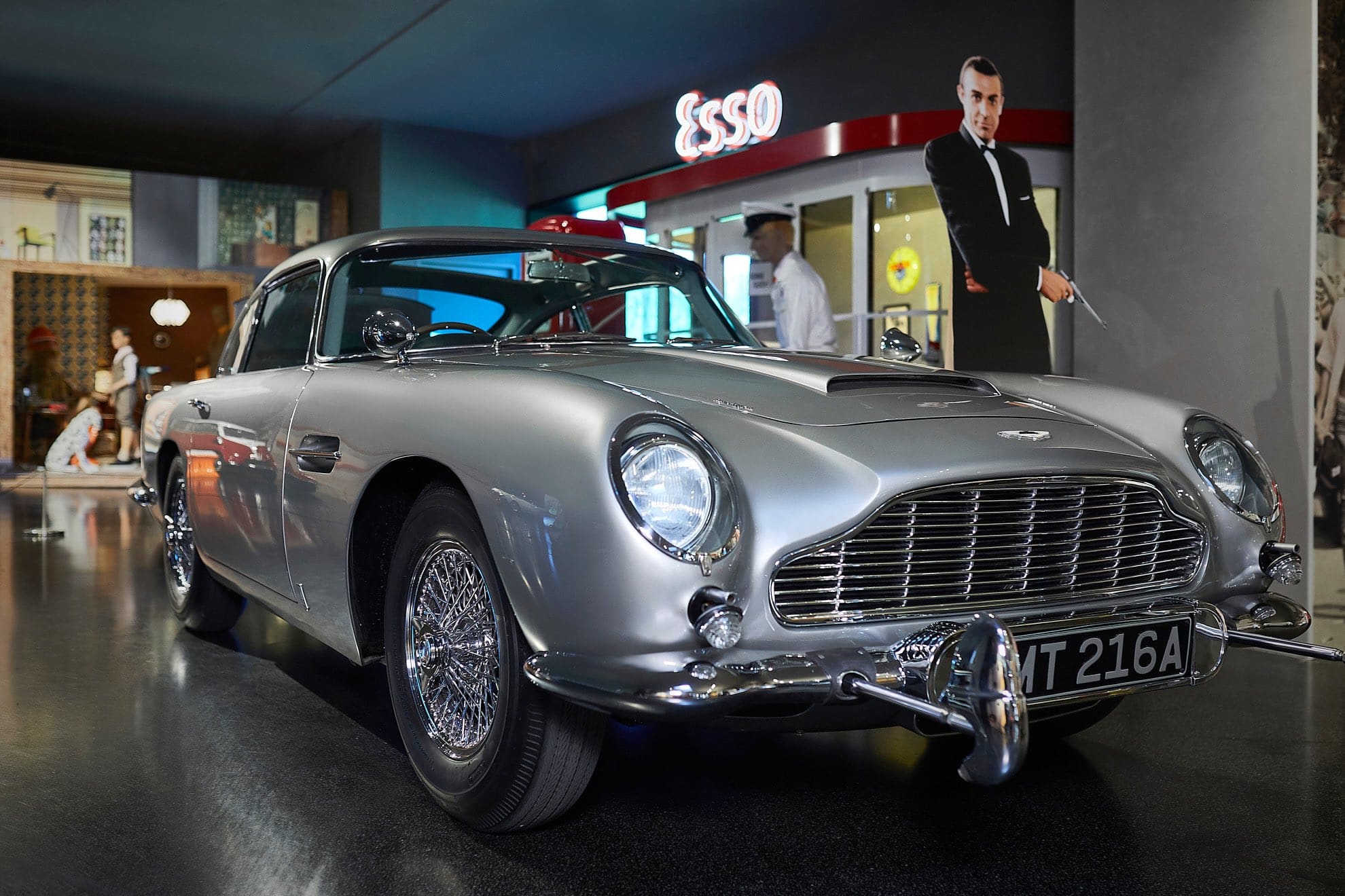 Im Innenbereich ist ein klassischer silberner Aston Martin-Sportwagen mit glänzenden Chromakzenten ausgestellt. Im Hintergrund steht ein Pappaufsteller eines Mannes im Anzug neben einem Esso-Schild und einem Interieur im Vintage-Stil.