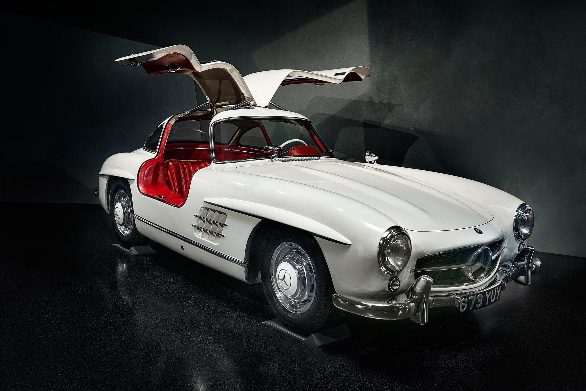 Ein klassischer weißer Mercedes-Benz 300SL mit markanten geöffneten Flügeltüren und leuchtend rotem Innenraum, ausgestellt in einem schwach beleuchteten Ausstellungsraum.