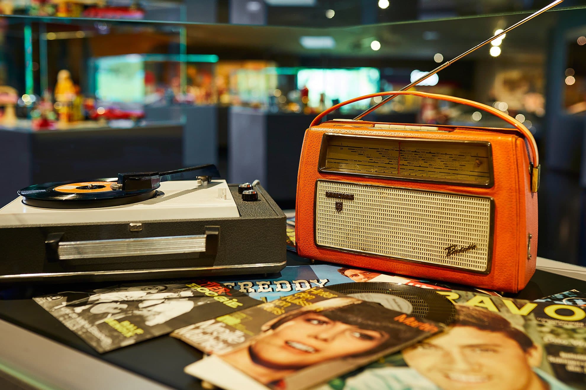 Auf einem Tisch in einem Retro-Ambiente liegen ein orangefarbenes Vintage-Radio, ein Plattenspieler mit einer Vinyl-Schallplatte und mehrere bunte Musikmagazine mit Prominenten-Gesichtern.