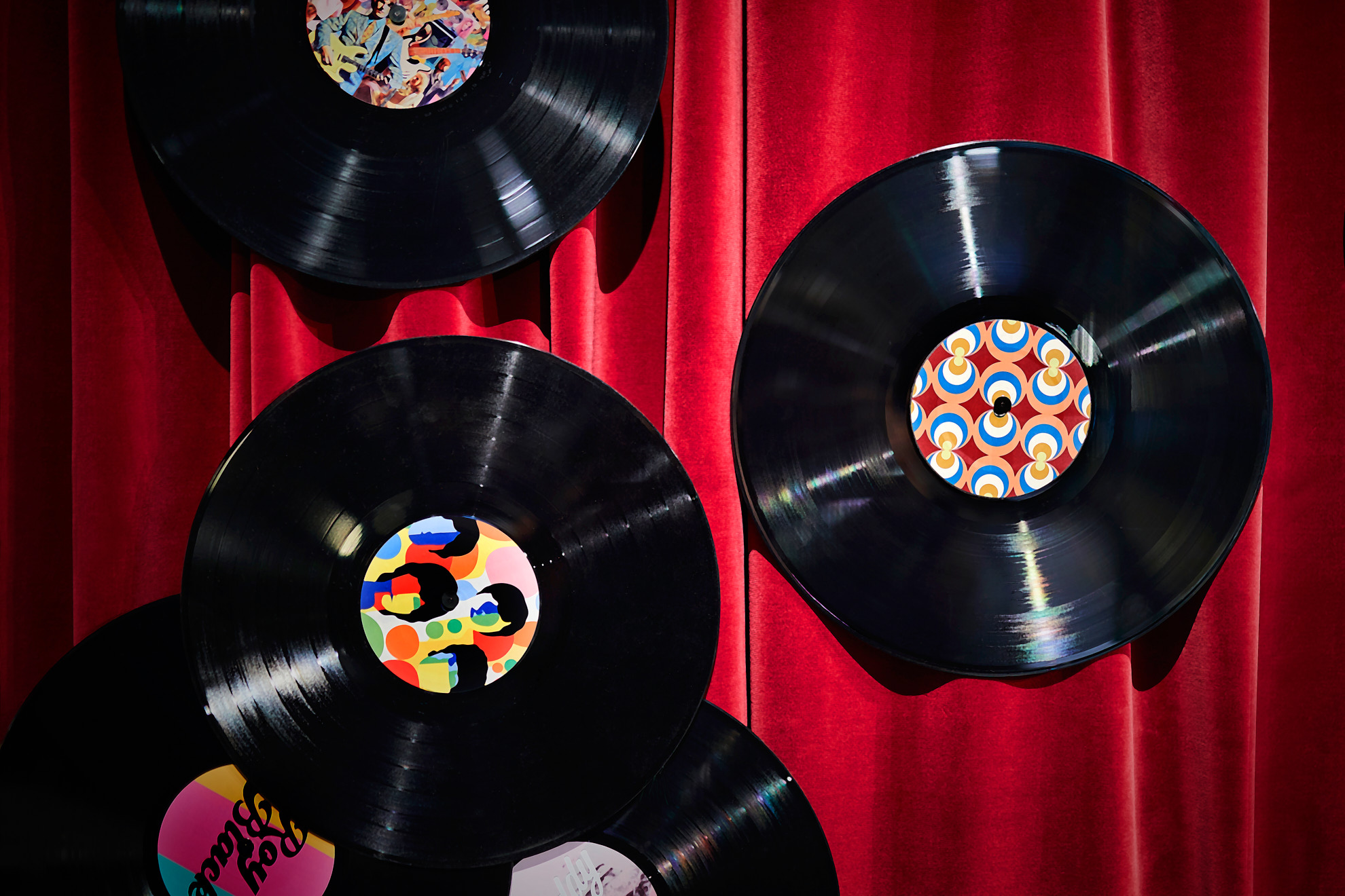 Vor einem Hintergrund aus rotem Samt werden vier Vinyl-Schallplatten mit bunten Etiketten präsentiert. Zwei Schallplatten überlappen sich teilweise, und eine ist in der unteren linken Ecke teilweise sichtbar.
