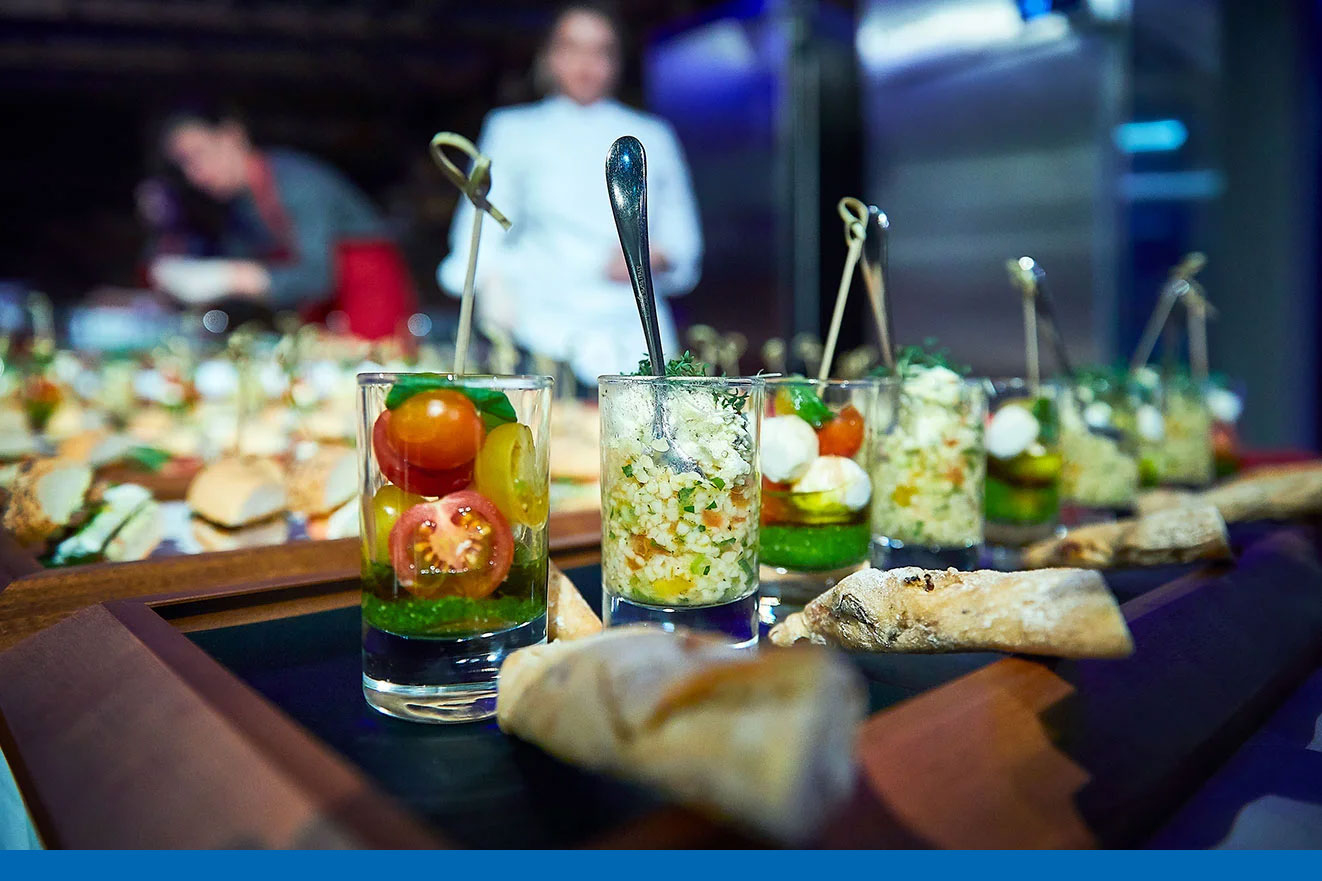 Fein angerichtete Vorspeisen im Glas mit Tomaten, Käse und Gemüse auf einem Tablett – im Hintergrund Küchenpersonal bei der Zubereitung