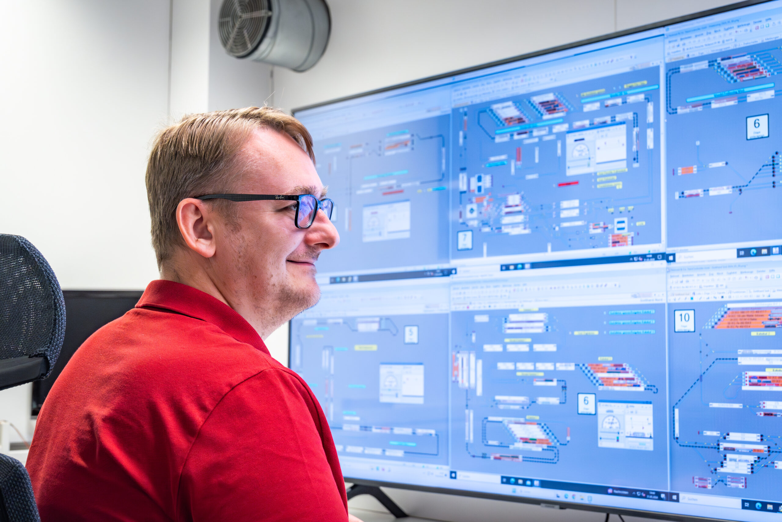 Ein Mann mit Brille und rotem Hemd sitzt vor großen Monitoren, auf denen komplexe Diagramme und Daten angezeigt werden. Möglicherweise überwacht oder steuert er ein technisches System in einer Büroumgebung.