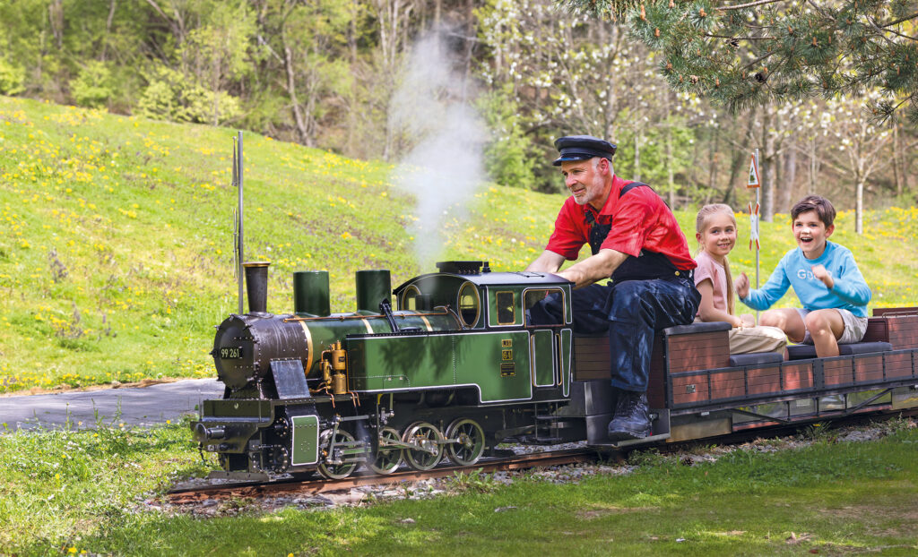 Ein als Zugführer verkleideter Mann fährt im Freien einen kleinen grünen Dampfzug, während hinter ihm zwei lächelnde Kinder in einem hölzernen Personenwagen durch einen grasbewachsenen, sonnenbeschienenen Park fahren.