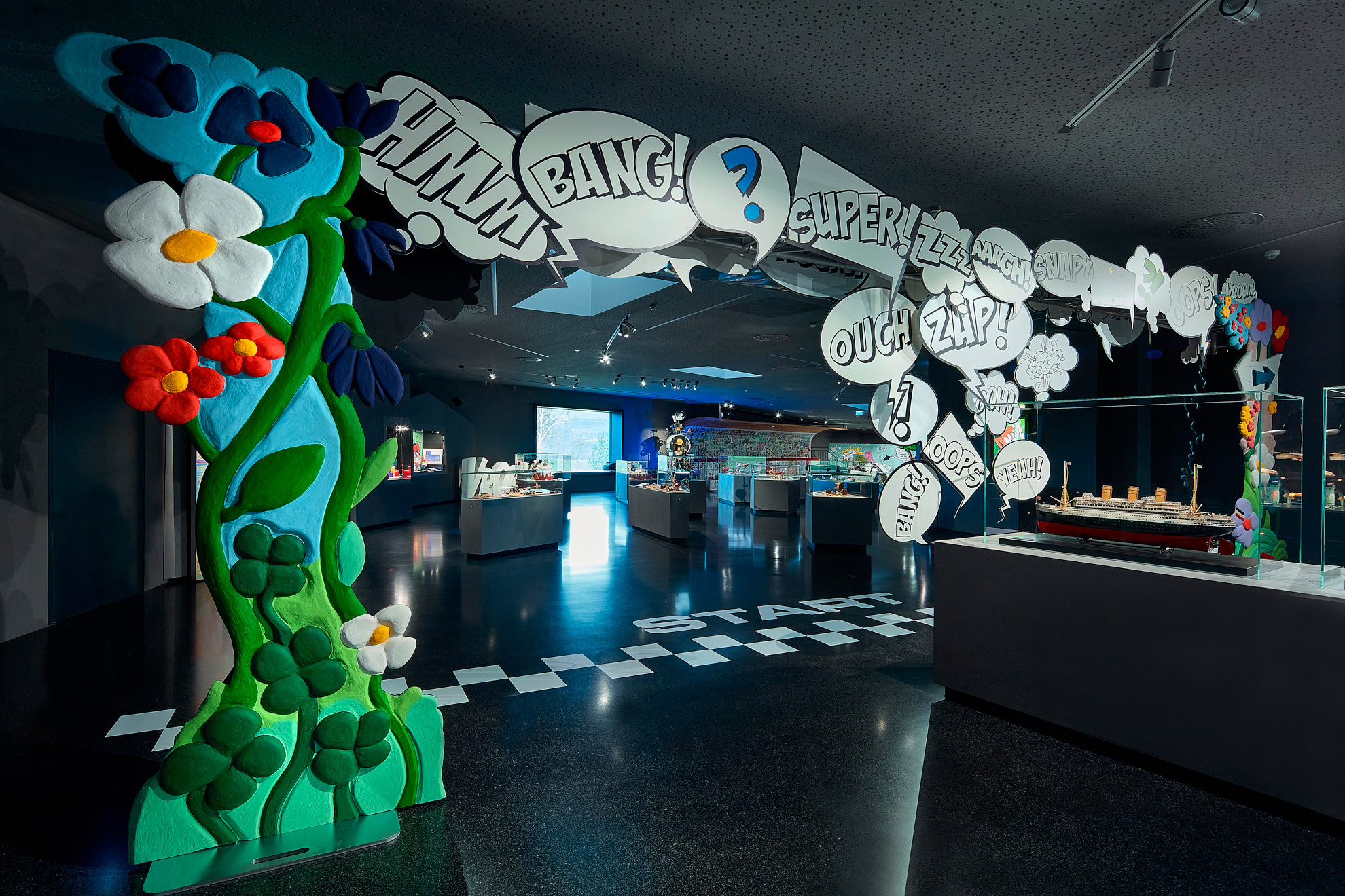 Comic-Display mit einem Zeichentricktier, Sprechblasen mit den Worten „VROOM!“ und „SPLASH!“ sowie an Schildern befestigten Federn. Auf der Oberfläche liegt ein gelbes Horn, im Hintergrund ist buntes Zeichentrick-Design zu sehen.