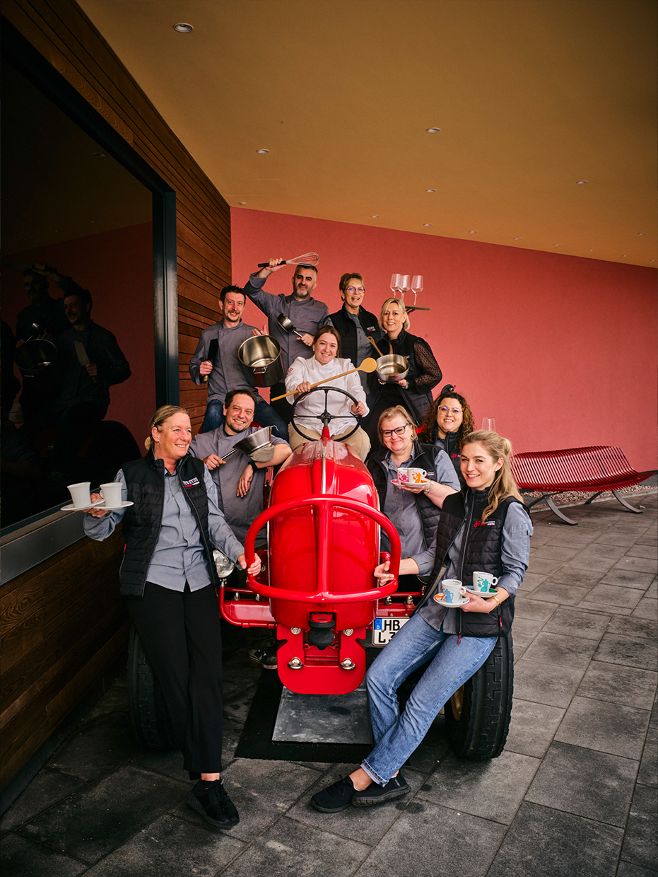 Eine Gruppe lächelnder Menschen in passenden grauen Hemden posiert mit Küchengeschirr um einen leuchtend roten Oldtimer-Traktor vor einem modernen Gebäude mit roten und braunen Wänden. Einige halten Tassen, Teller, Pfannen oder Weingläser.