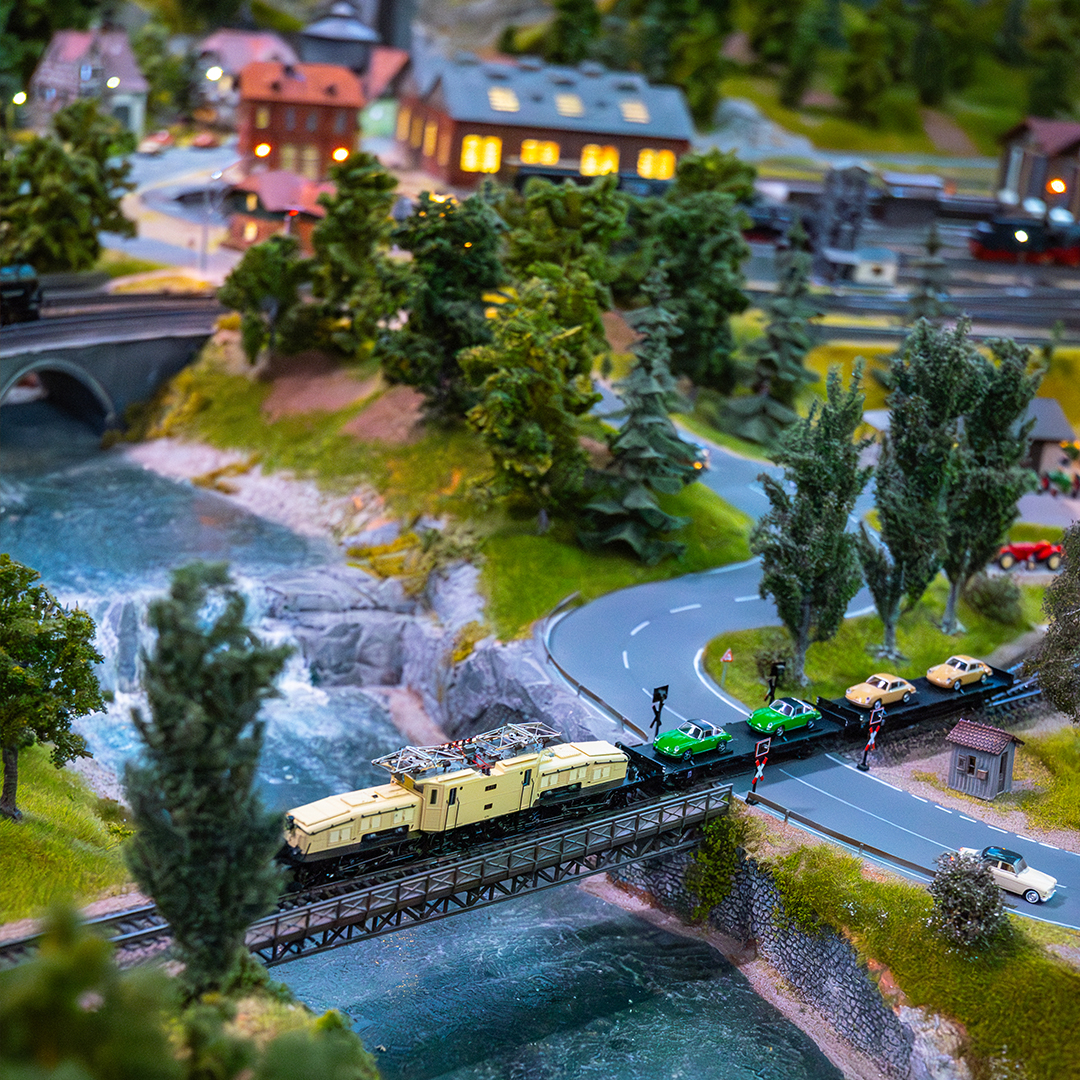 Eine detaillierte Modelleisenbahnszene zeigt eine gelbe Lokomotive, die eine Brücke über einen Fluss überquert, gewundene Straßen mit Autos, üppige Bäume und beleuchtete Gebäude im Hintergrund, die eine lebendige Miniaturlandschaft schaffen.