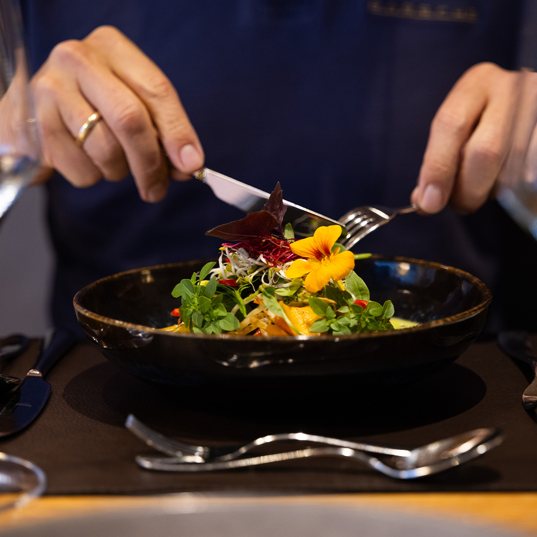 Eine Person im blauen Hemd isst mit Messer und Gabel einen bunten Gourmetsalat, garniert mit essbaren Blüten und Microgreens, der in einer schwarzen Schüssel auf einem gedeckten Esstisch serviert wird.