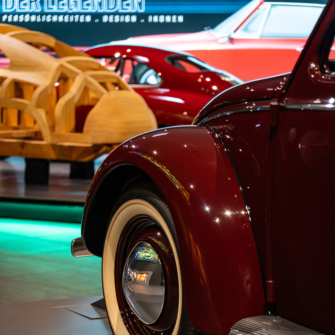 Nahaufnahme des vorderen Kotflügels und des Weißwandreifens eines roten Oldtimers, im Hintergrund sind auf einer Autoausstellung ein weiterer roter Oldtimer und ein hölzerner Autorahmen zu sehen.