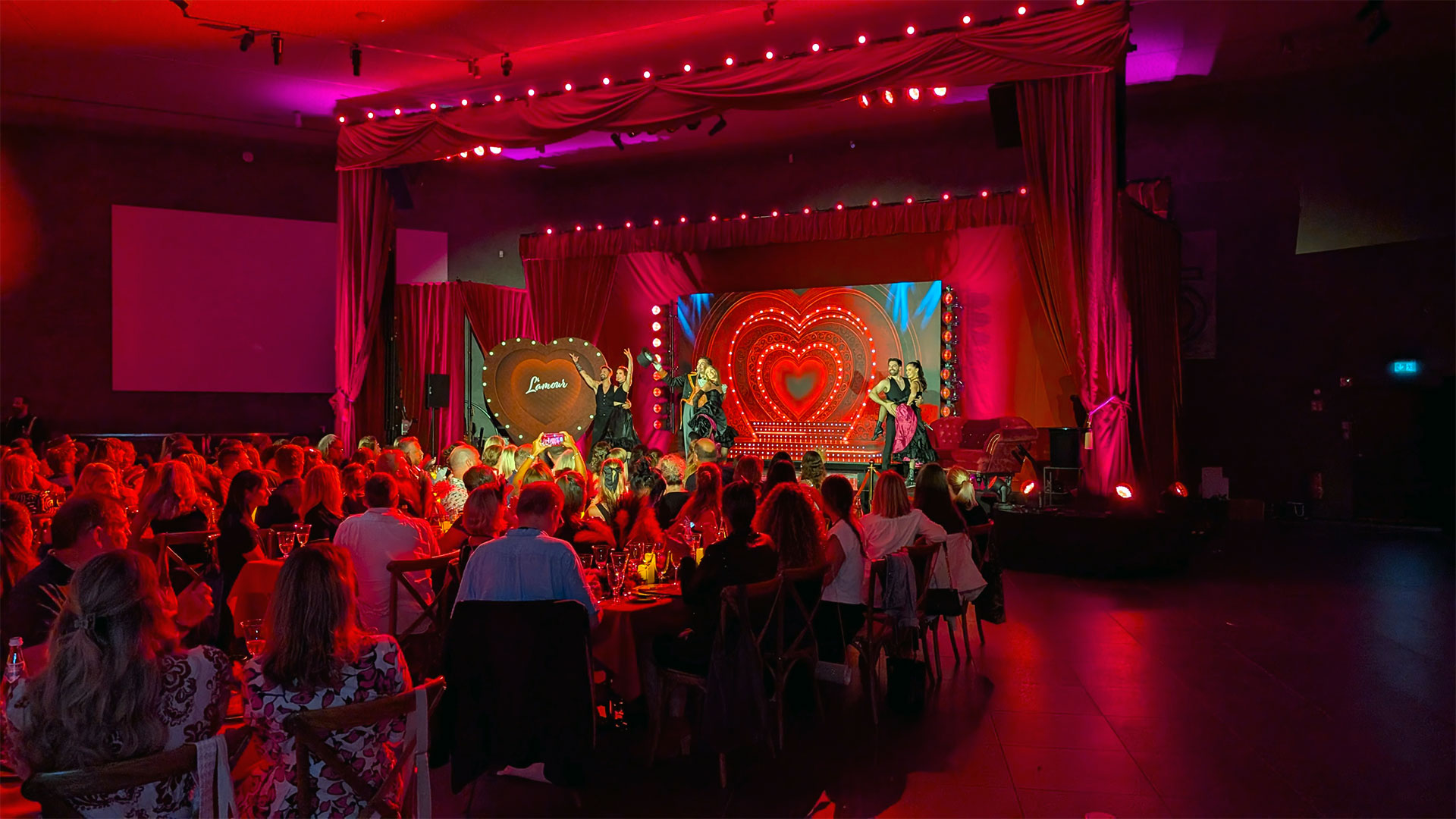 Ein lebhaftes Publikum verfolgt eine Bühnenshow mit Darstellern in leuchtenden Kostümen, bunten Lichtern und herzförmigen Dekorationen bei rosa und roter Beleuchtung in einem dekorierten Theater.