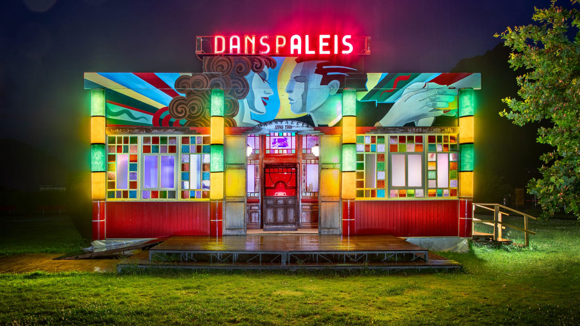 Ein farbenfrohes, nachts beleuchtetes Gebäude mit dem Neonschild DANSPALEIS an der Spitze, mit Buntglasfenstern und einer Wandmalerei mit stilisierten Gesichtern und Händen an der Fassade. Eine kleine Holzrampe führt zum Eingang.