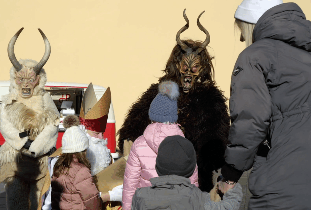 Zwei als gehörnte Krampusmonster verkleidete Personen interagieren an einem sonnigen Tag draußen mit Kindern und einem Erwachsenen. Die Kinder tragen Winterjacken und gestrickte Mützen. Eine weitere Person in einem festlichen Kostüm ist teilweise sichtbar.