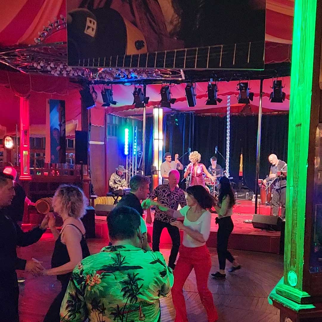 Menschen tanzen energisch vor einer Live-Band, die auf einer farbenfrohen, gut beleuchteten Bühne in einem lebhaften Lokal mit roten Vorhängen und Holzfußboden auftritt. Helle grüne und blaue Lichter erhellen die Szene.
