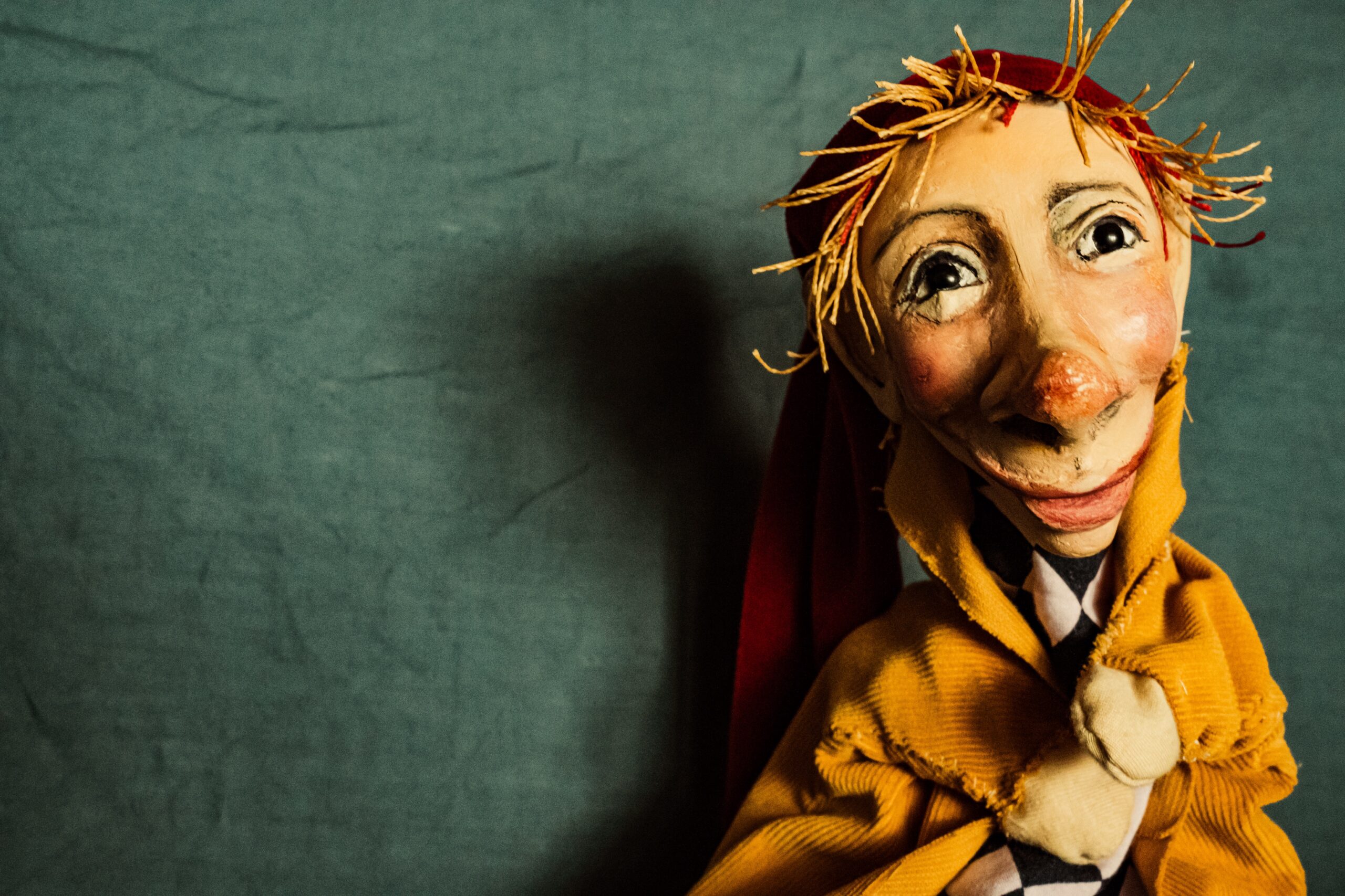 Eine handgefertigte Puppe mit großer Nase, ausdrucksstarken Augen und strohigem, gelbem Haar trägt einen gelben Mantel und eine rote Kapuze vor einem schlichten dunkelgrünen Hintergrund.