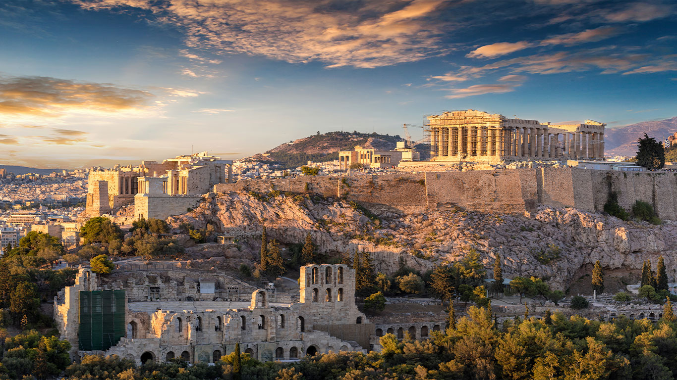 Die Akropolis von Athen bei Sonnenuntergang, mit dem Parthenon und anderen antiken Ruinen auf einem felsigen Hügel, der die Stadt überragt und von Grün und fernen Bergen umgeben ist.