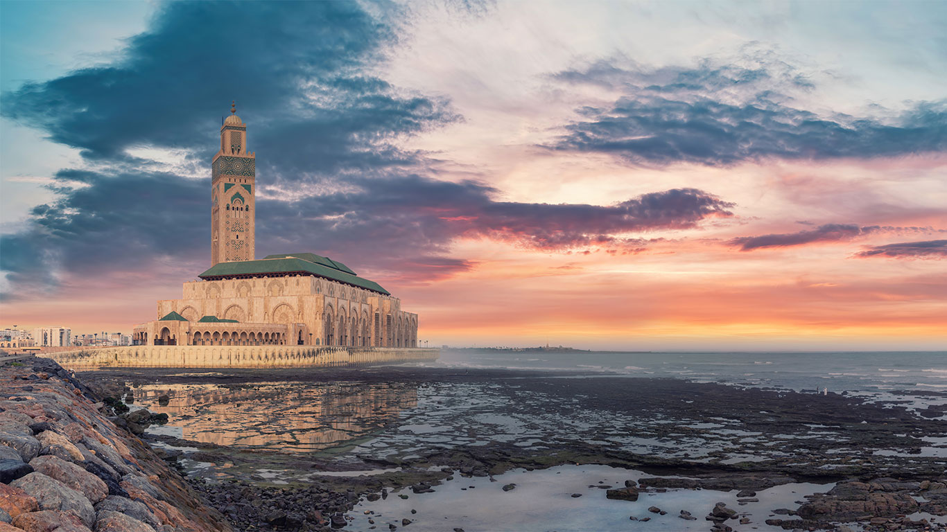Eine große Moschee mit einem hohen Minarett steht bei Sonnenuntergang direkt am Meer und spiegelt sich im seichten Wasser. Der Himmel ist mit leuchtenden Wolken in Rosa-, Lila- und Orangetönen gefüllt. Im Vordergrund sind felsige Küstenlinien zu sehen.
