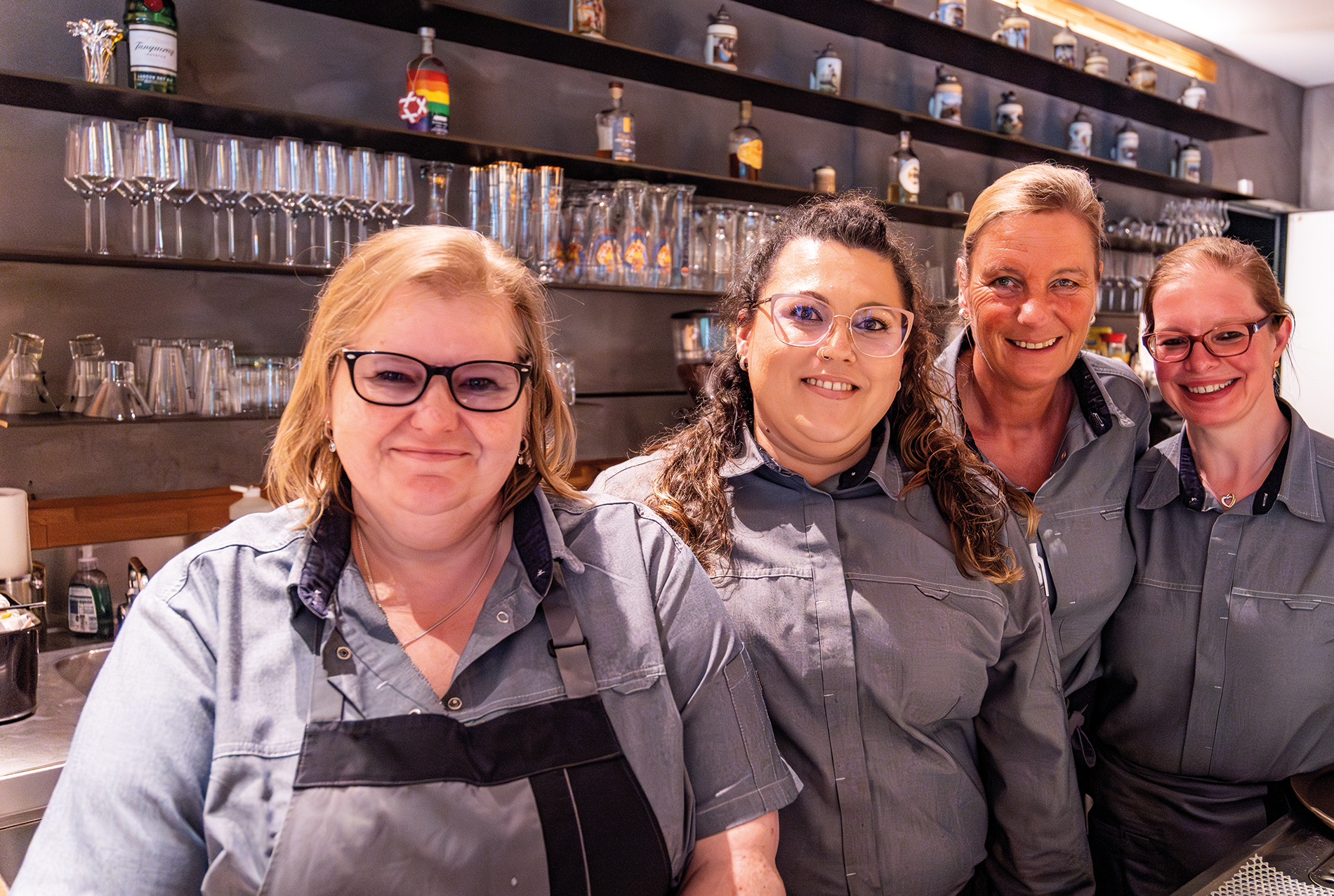 Vier lächelnde Frauen in passenden grauen Uniformen stehen hinter einer Bar mit Regalen voller Gläser und Flaschen im Hintergrund. Sie scheinen Mitarbeiter zu sein, die in einer freundlichen, gut beleuchteten Umgebung zusammen posieren.