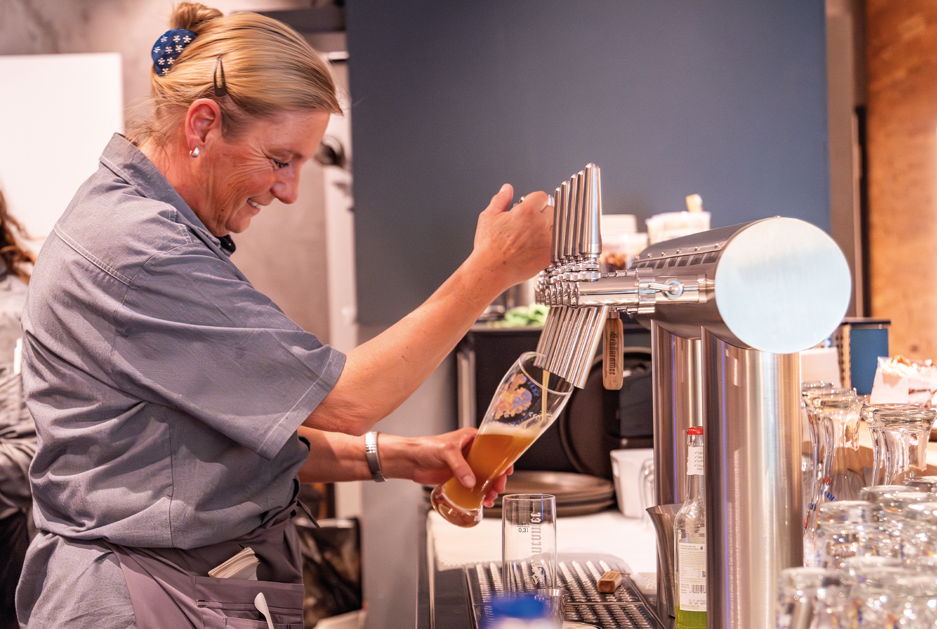 Eine lächelnde Frau in einem grauen Hemd gießt Bier aus einem Zapfhahn in ein Glas an einer Bar, auf deren Tresen saubere Gläser und Barausstattung zu sehen sind.