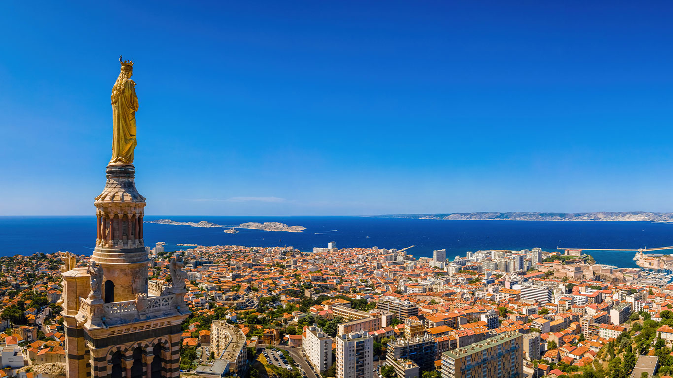 Eine goldene Statue auf einem Kirchturm überblickt eine Küstenstadt mit orangefarbenen Dächern, blauem Meer und entfernten Inseln unter einem klaren, hellen Himmel.
