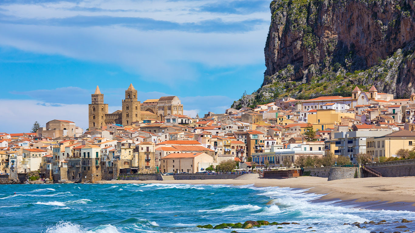 Buntes Küstenstädtchen mit terrakottabedeckten Gebäuden und einer großen Kirche vor einer felsigen Klippe. Blaue Wellen schlagen an den Sandstrand im Vordergrund unter einem teilweise bewölkten Himmel.