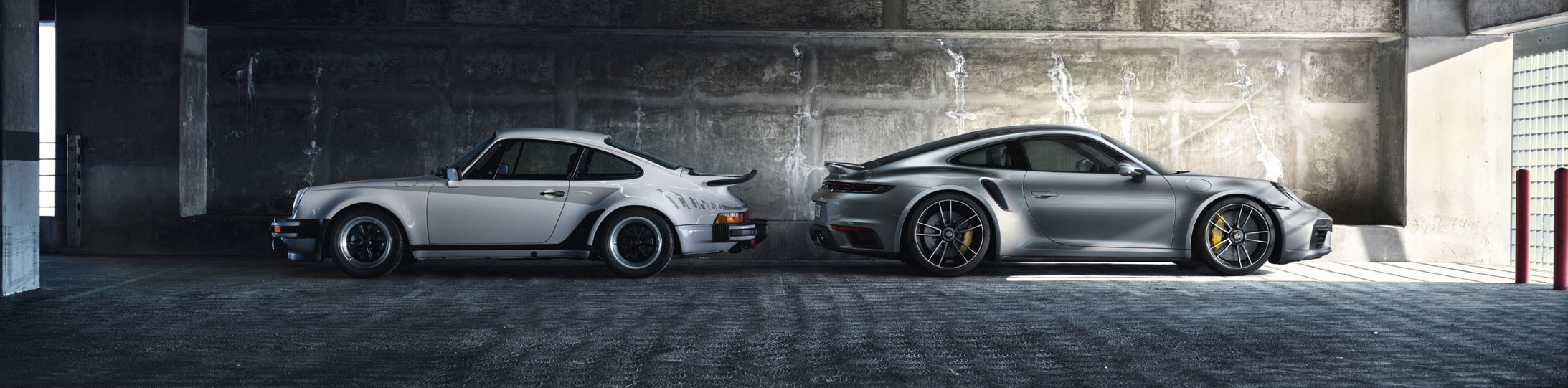 Ein klassischer weißer Porsche 911 Turbo parkt neben einem modernen silbernen Porsche 911 Turbo in einer industriellen Betongarage, beide im Profil gezeigt.