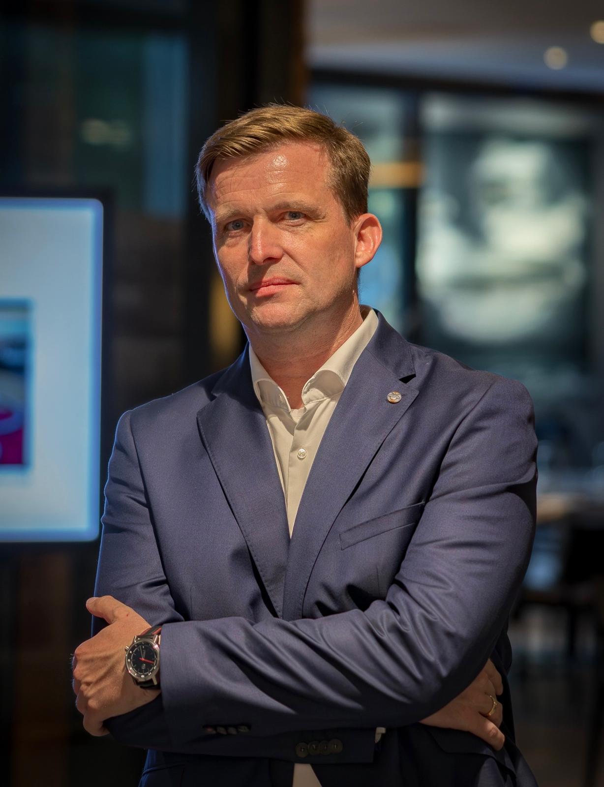 Sven Bössow in blauem Anzug und weißem Hemd steht mit verschränkten Armen in einem Haus und schaut in die Kamera. Der Hintergrund ist leicht unscharf und zeigt eine moderne, professionelle Umgebung mit großen Fenstern.