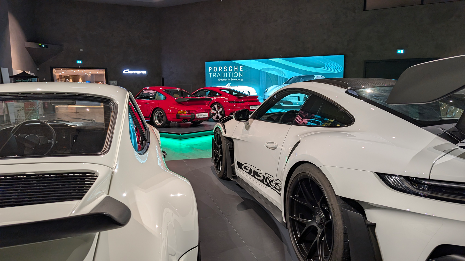 Ein Ausstellungsraum mit mehreren ausgestellten Porsche Sportwagen, darunter weiße und rote Modelle. Im Hintergrund ist ein großes beleuchtetes Porsche Tradition-Schild zu sehen. Der Raum ist modern und schlank eingerichtet.