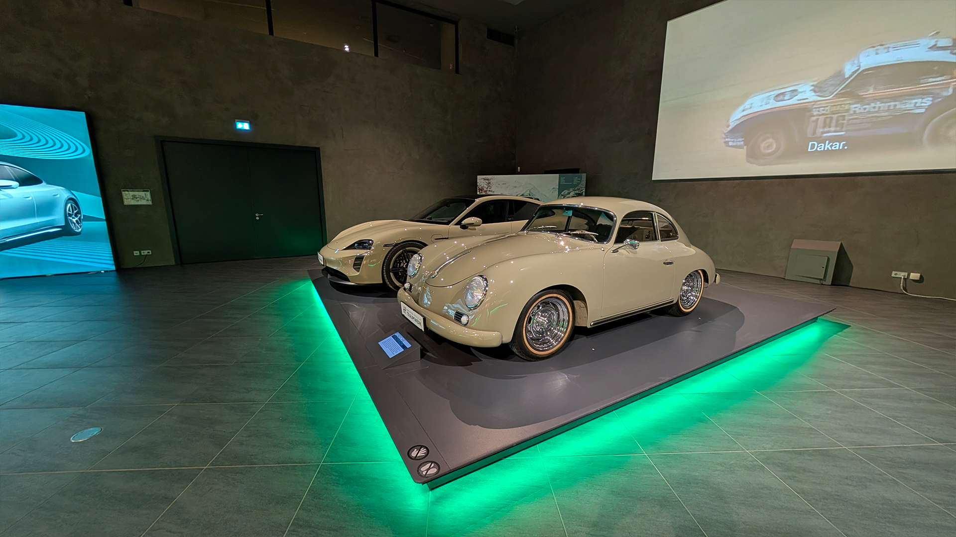 Zwei klassische weiße Porsche stehen auf einer erhöhten Plattform mit grüner Beleuchtung in einem modernen, schwach beleuchteten Ausstellungsraum. Im Hintergrund sind ein projiziertes Rennvideo und andere Displays zu sehen.