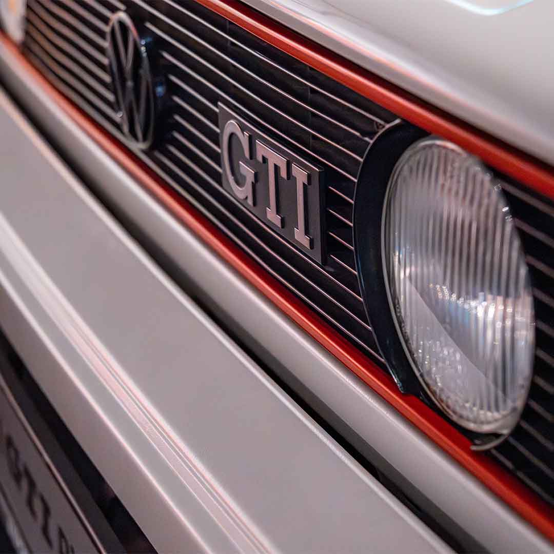 Nahaufnahme des vorderen Kühlergrills und der Scheinwerfer eines Volkswagens mit GTI-Plakette und VW-Emblem, mit schwarzen horizontalen Lamellen und einem roten Zierstreifen.