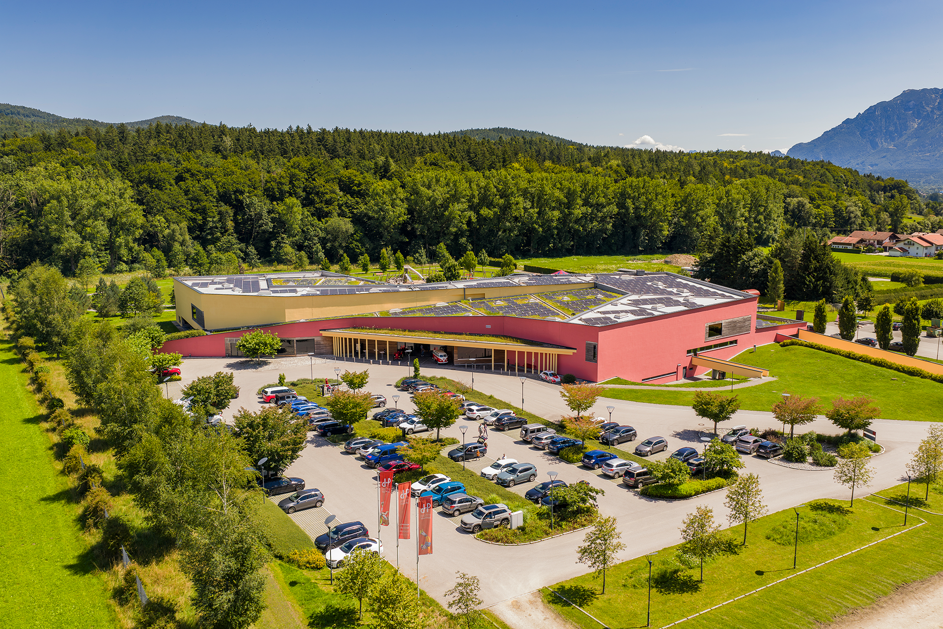 Ein großes, modernes Gebäude mit roten Außenwänden steht inmitten von grünen Bäumen und Hügeln. Davor befindet sich ein Parkplatz mit vielen Autos, und im Hintergrund sind der blaue Himmel und die Berge zu sehen.
