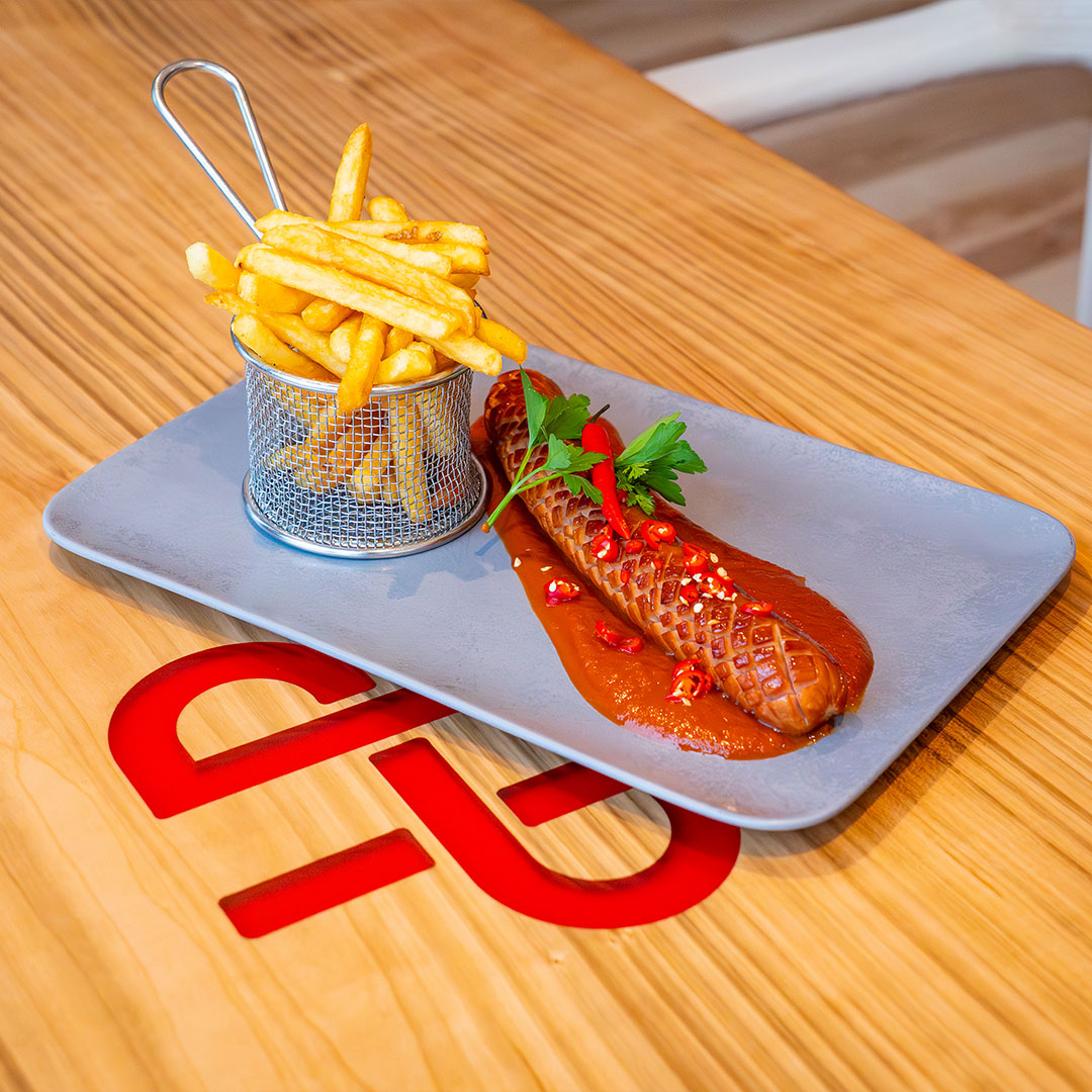 Auf einem grauen Teller auf einem Holztisch steht ein Metallkorb mit Pommes frites und einer gegrillten Wurst in scharfer roter Soße, garniert mit gehacktem Chili und Petersilie. Der Tisch ist mit einem roten Logo unter dem Teller versehen.