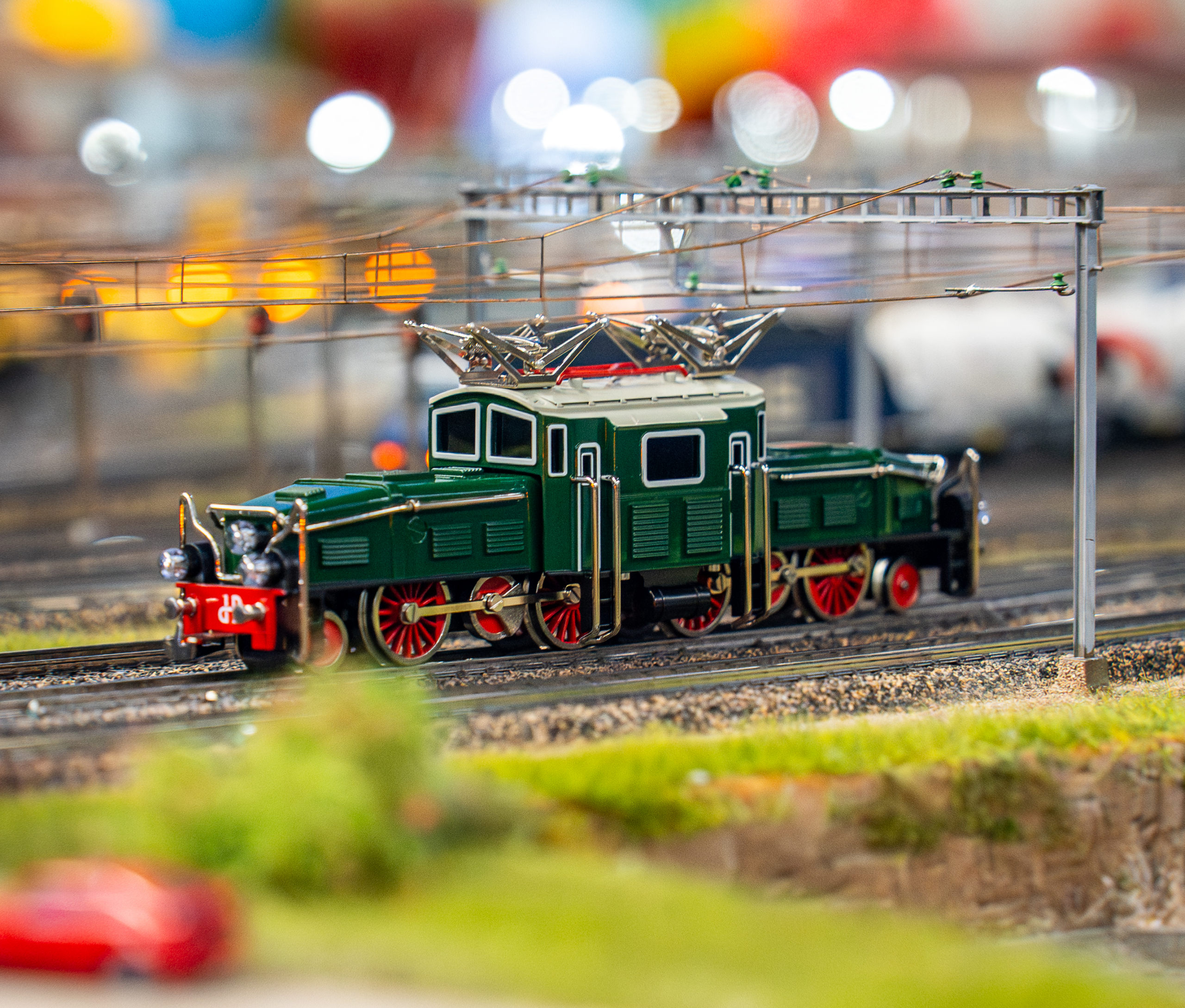 Ein detailliertes Modell eines grünen Elektrozuges mit roten Rädern steht auf Miniaturgleisen, umgeben von verschwommenen Hintergrundelementen und bunten Lichtern.