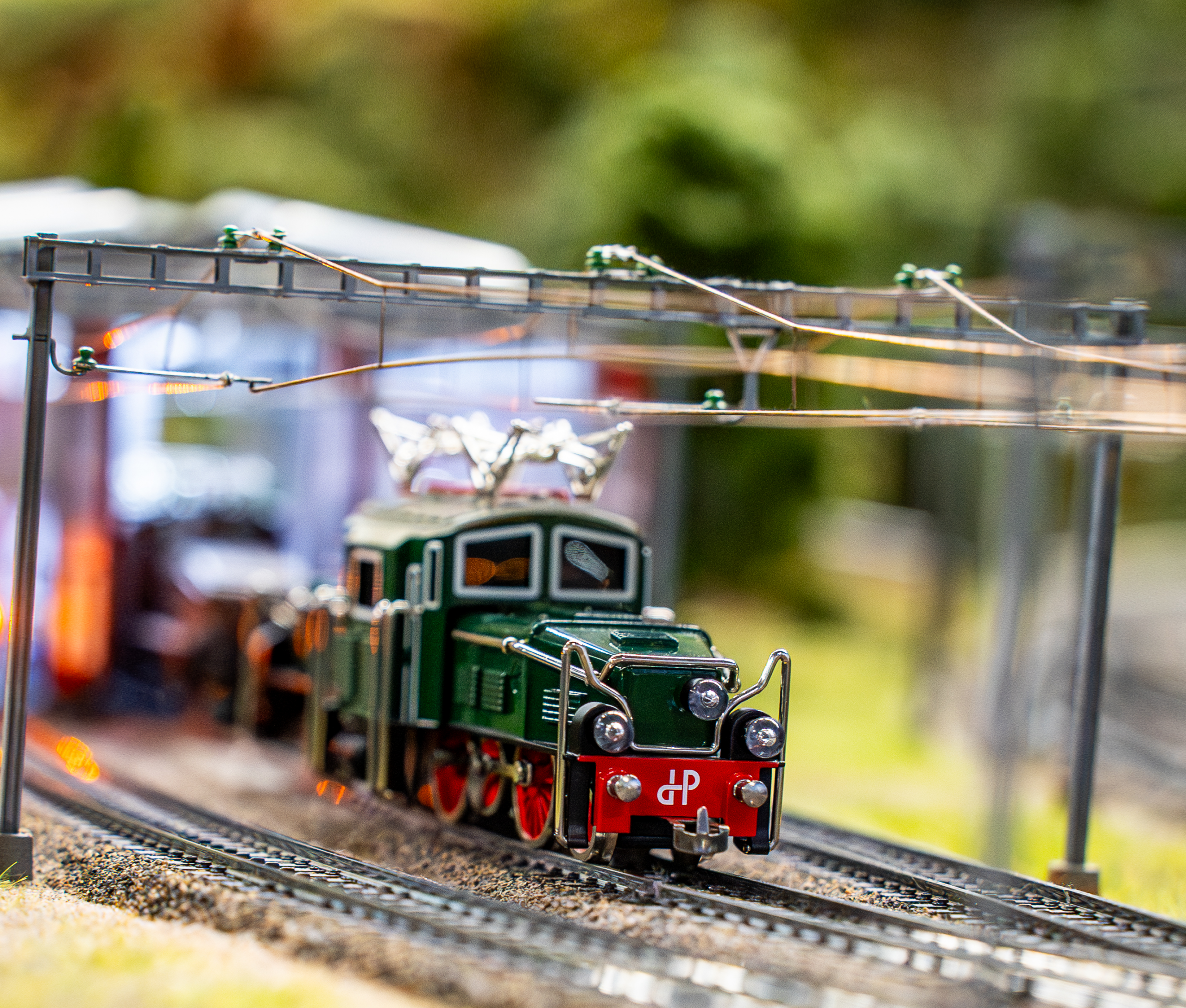 Ein detaillierter Modellzug mit grüner Lokomotive und roten Akzenten fährt auf Miniaturgleisen unter Oberleitungen, umgeben von verschwommenem Grün und Strukturen im Hintergrund.